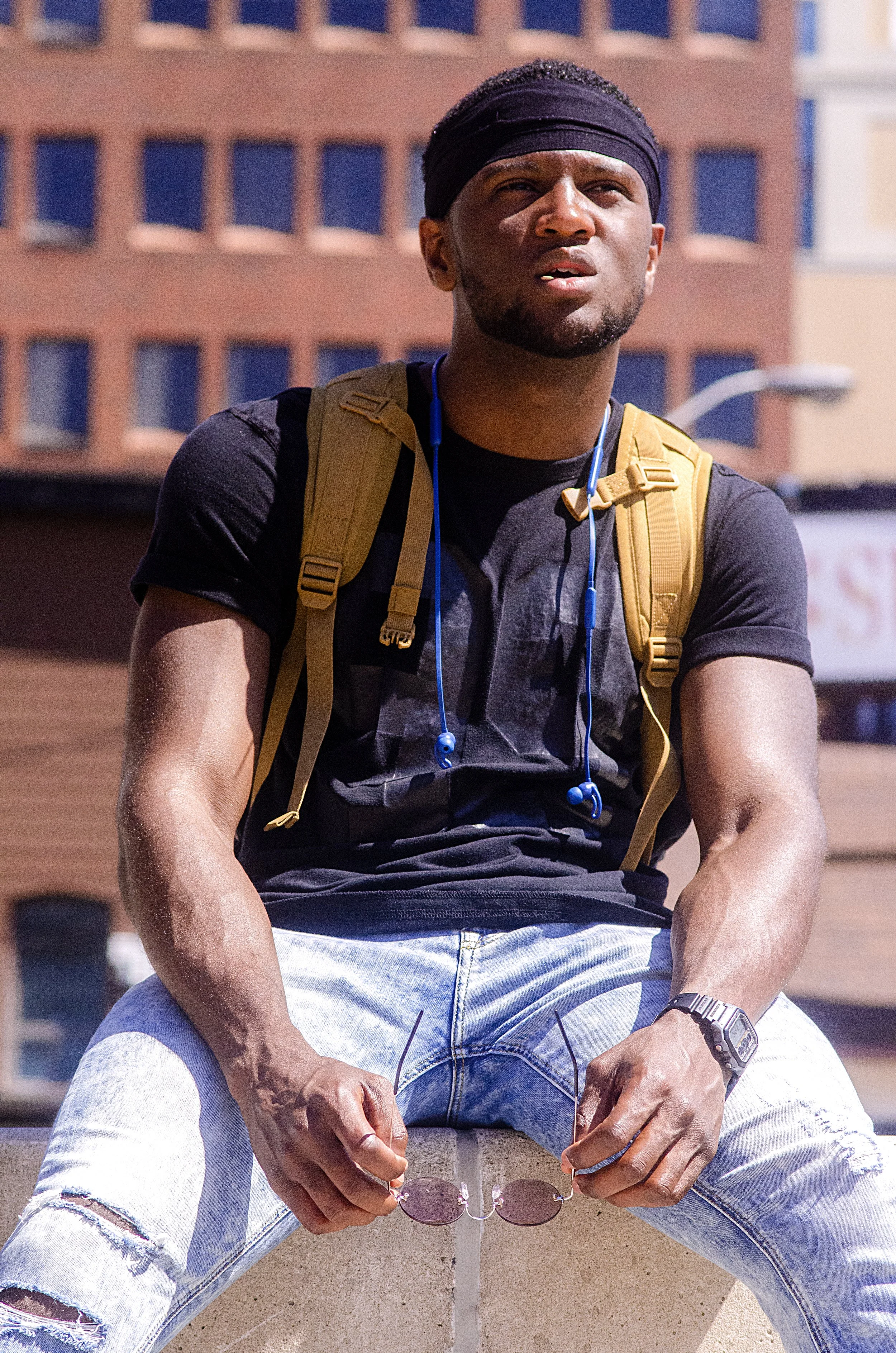 A man sits on a ledge with his legs hanging over the edge, holding a pair of sunglasses and looking upward while squinting in the bright sunlight. He is wearing a black shirt, blue jeans, a black headband, and a brown backpack.
