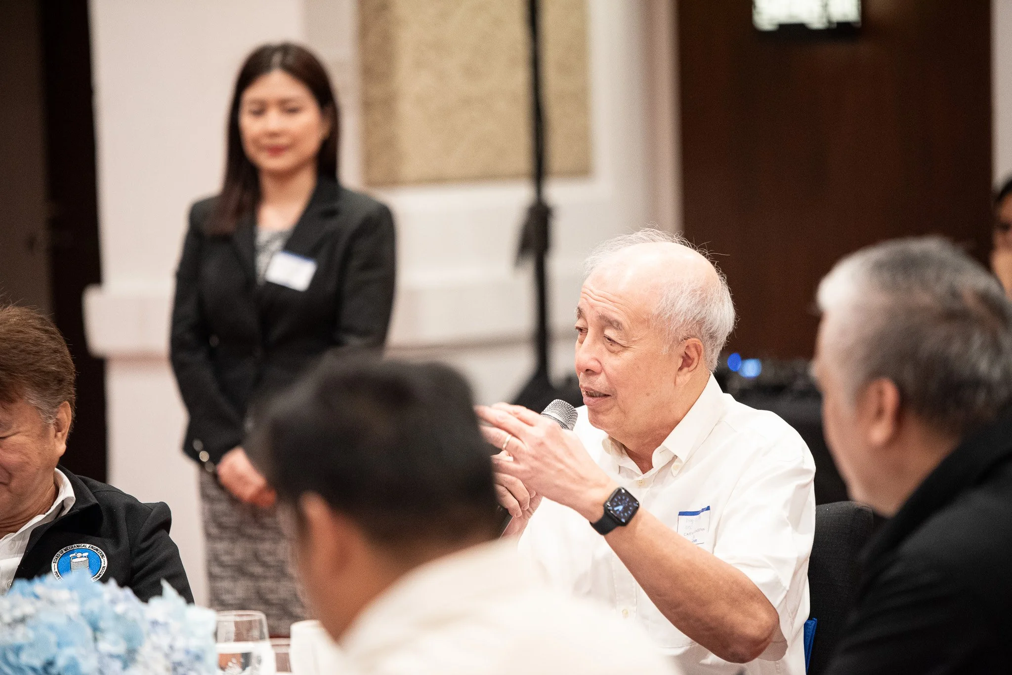An elderly man with grey hair speaking into a microphone at a formal gathering, with other people listening. A woman in a black blazer stands in the background.