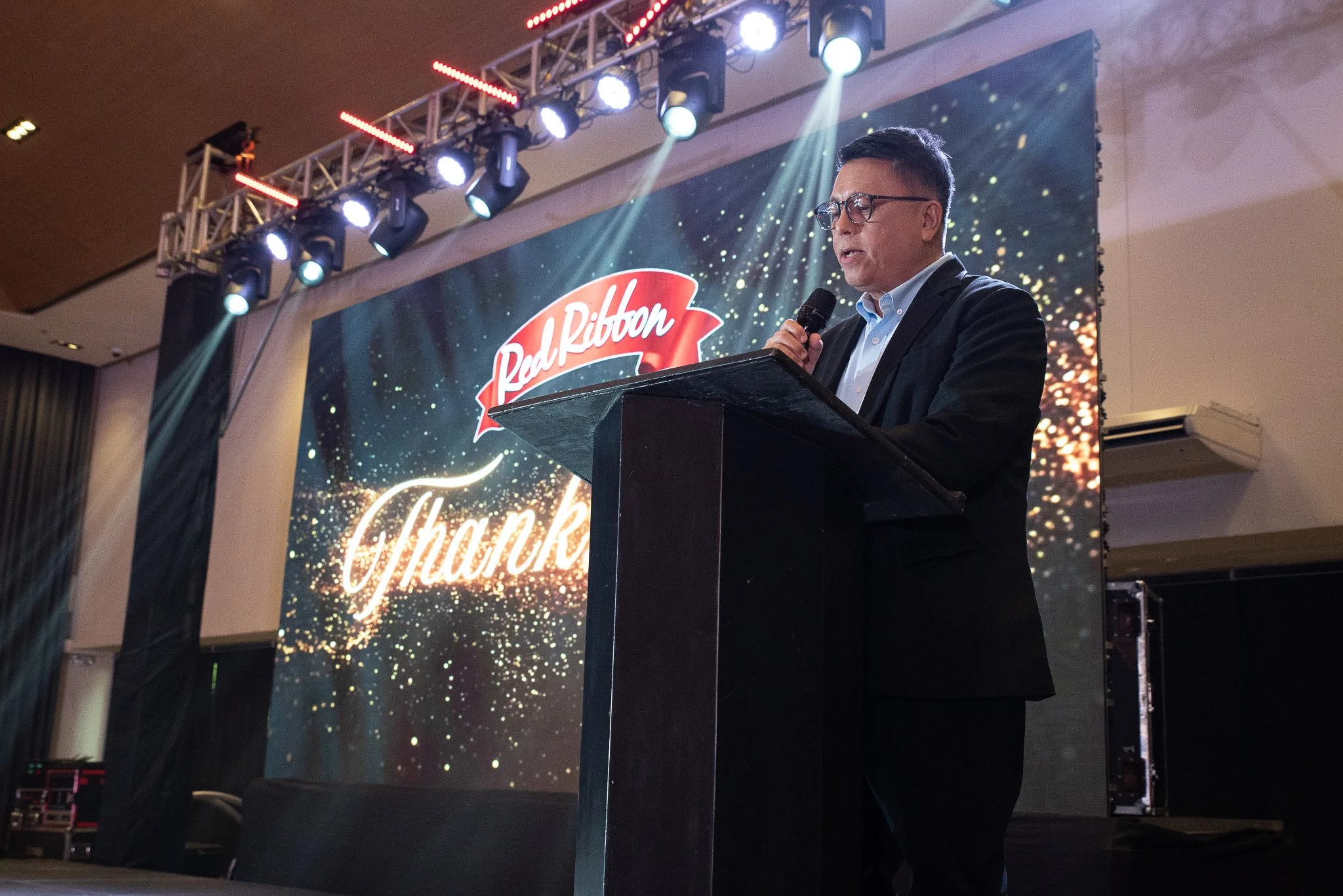 A man in a black suit and glasses speaks at a podium during an event. Behind him is a large screen displaying the Red Ribbon logo and the word "Thank" with confetti and sparkles.