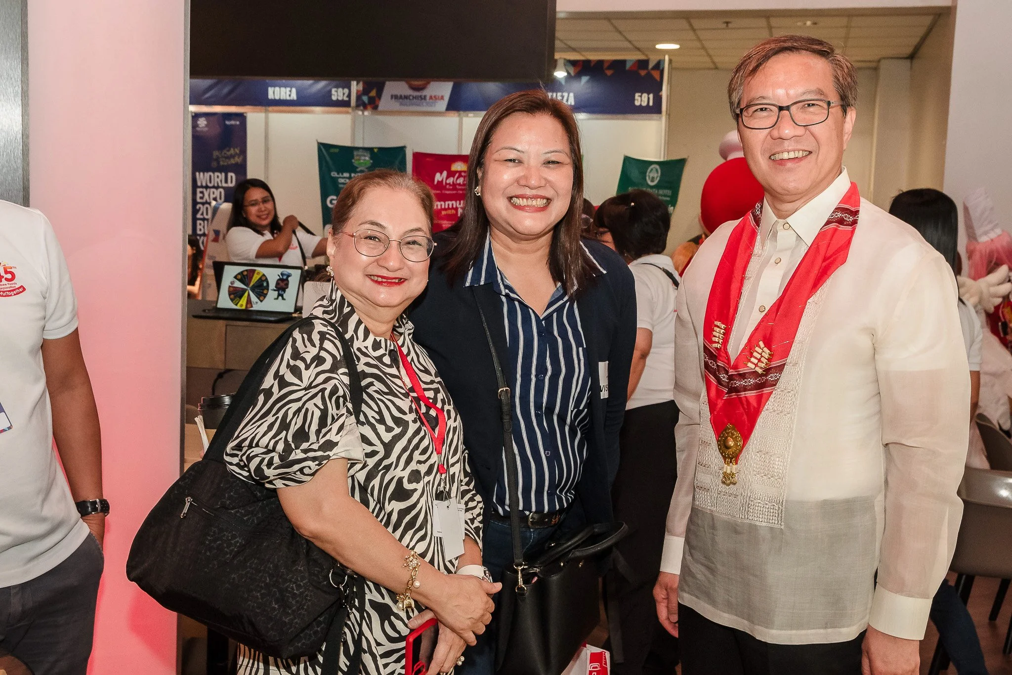 Three smiling people, two women and one man, standing together at an indoor event, with booths and banners in the background.