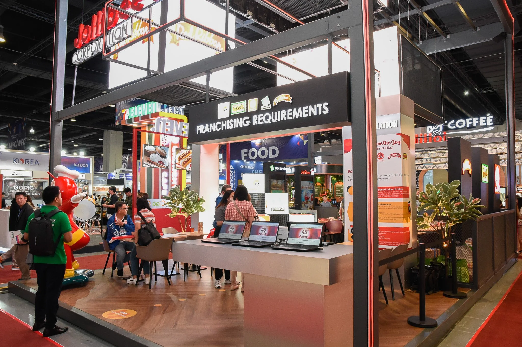 Trade show booth with a sign that reads 'Franchising Requirements' and several laptops on display in the foreground. People are walking and sitting inside the booth, with bright lights and colorful signage around the exhibition hall.