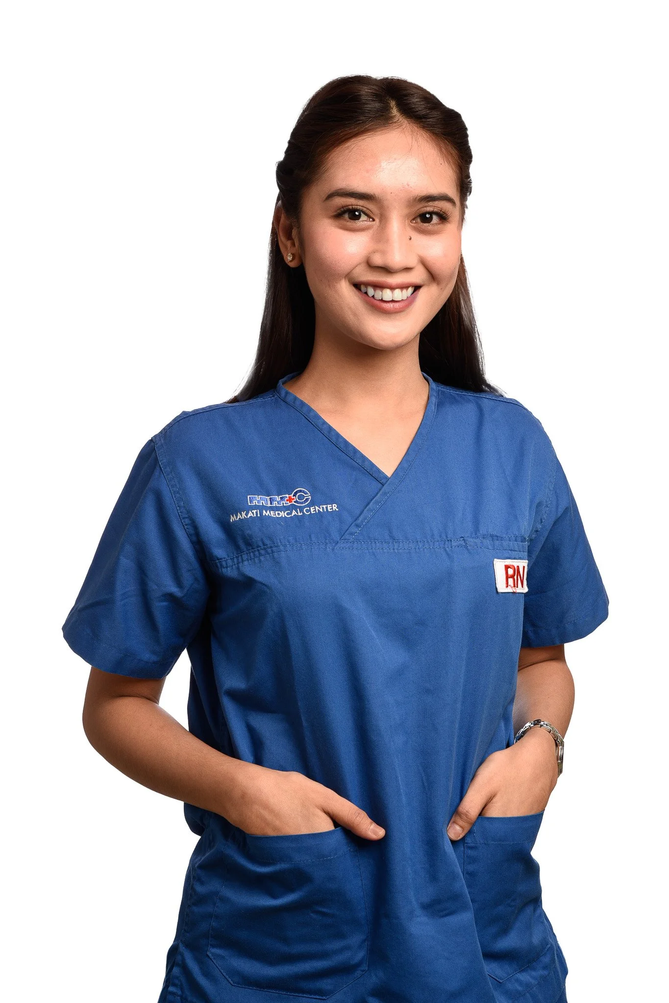 Young woman wearing blue medical scrubs with a smile, hands in pockets, standing against a white background.