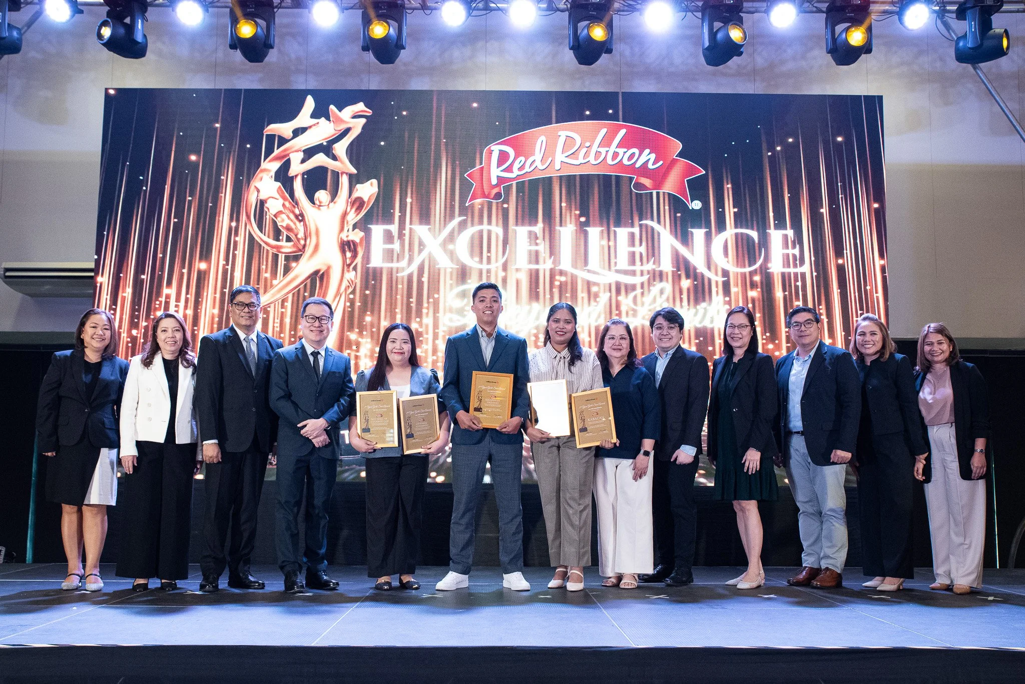 Group of people standing on stage during an awards ceremony with a large digital display behind them featuring the Red Ribbon Excellence logo.