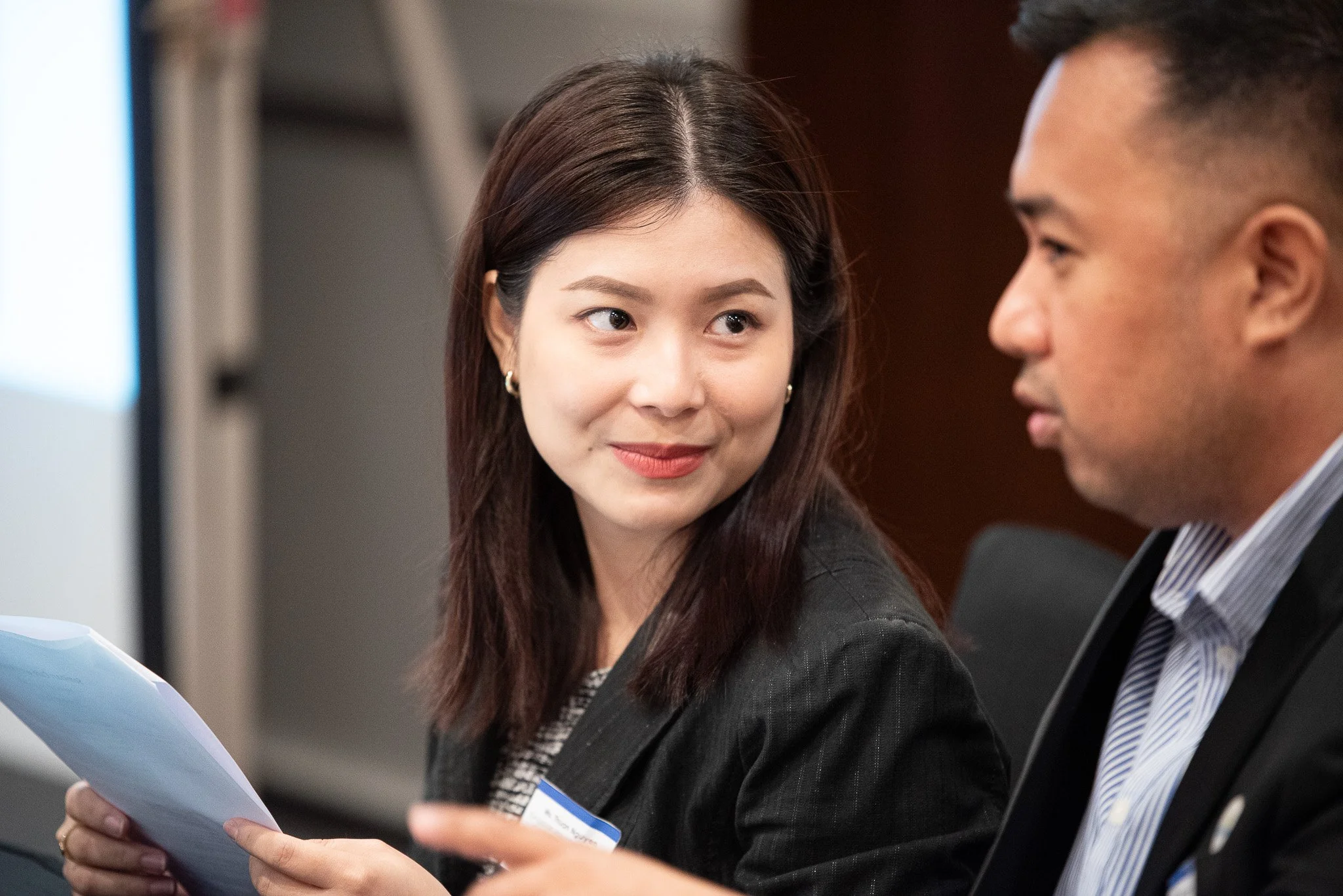 A woman and a man sitting and talking during a business meeting, with the woman holding papers and looking at the man.