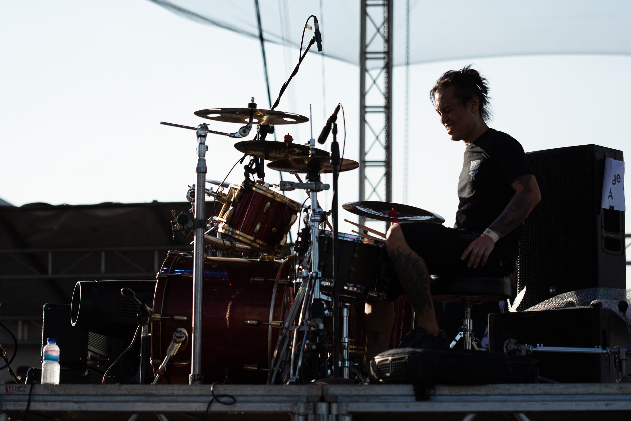 A person playing the drums on an outdoor stage with stage lights and metal framework in the background, silhouetted against bright sky.