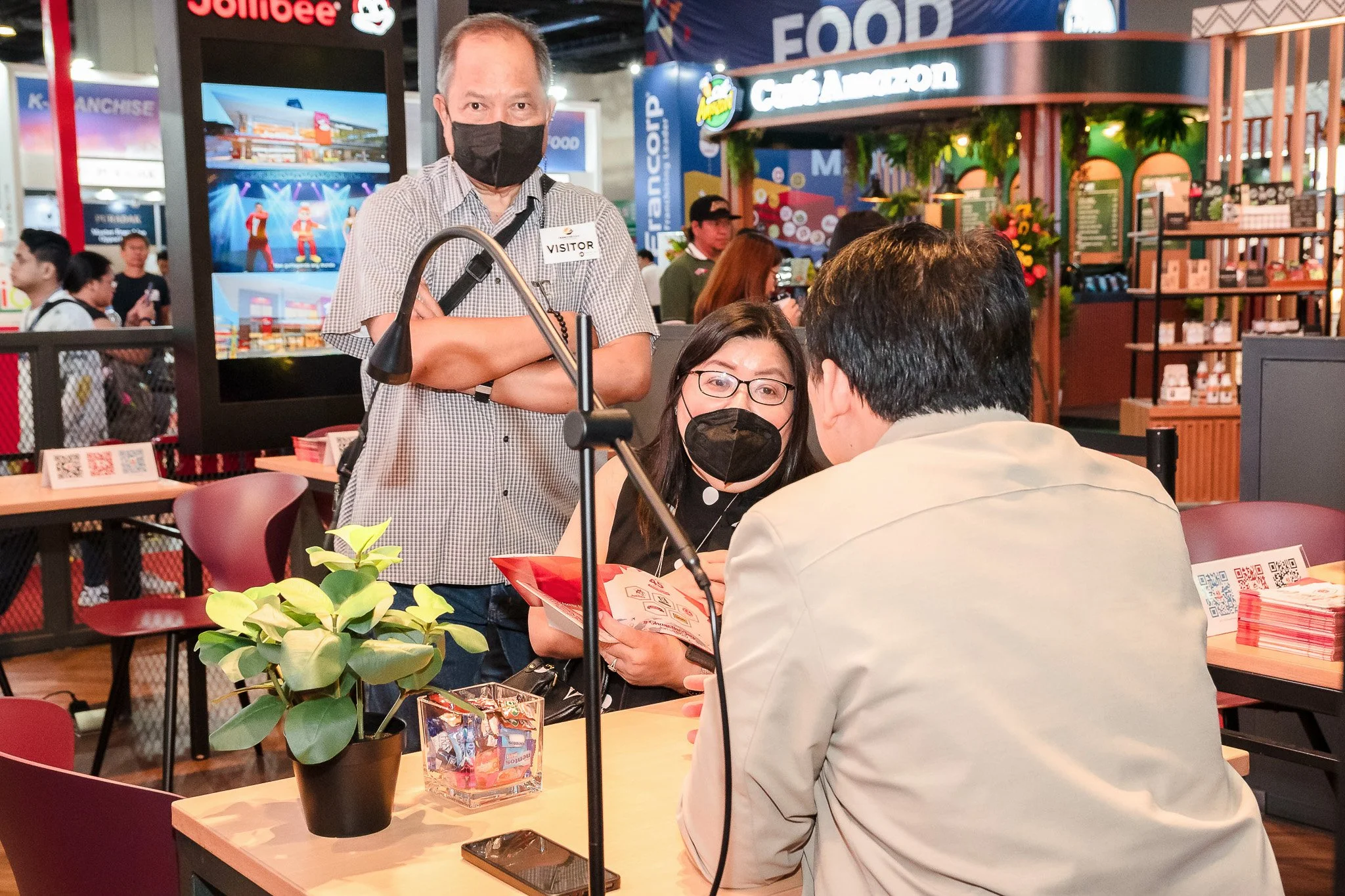 Three people wearing masks engaged in a discussion at a food event or trade show. One woman is holding a booklet, while a man with his back to the camera listens. A man standing nearby, also masked, observes the conversation. The table has a potted p