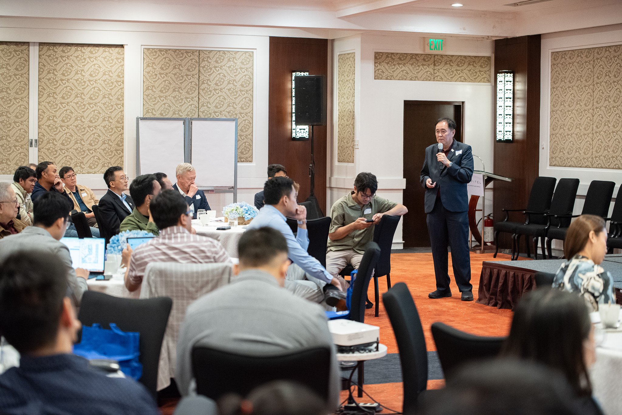 A man in a dark suit is giving a presentation with a microphone in a conference room filled with attendees seated at tables. The room has beige patterned wallpaper and an exit sign above the door.