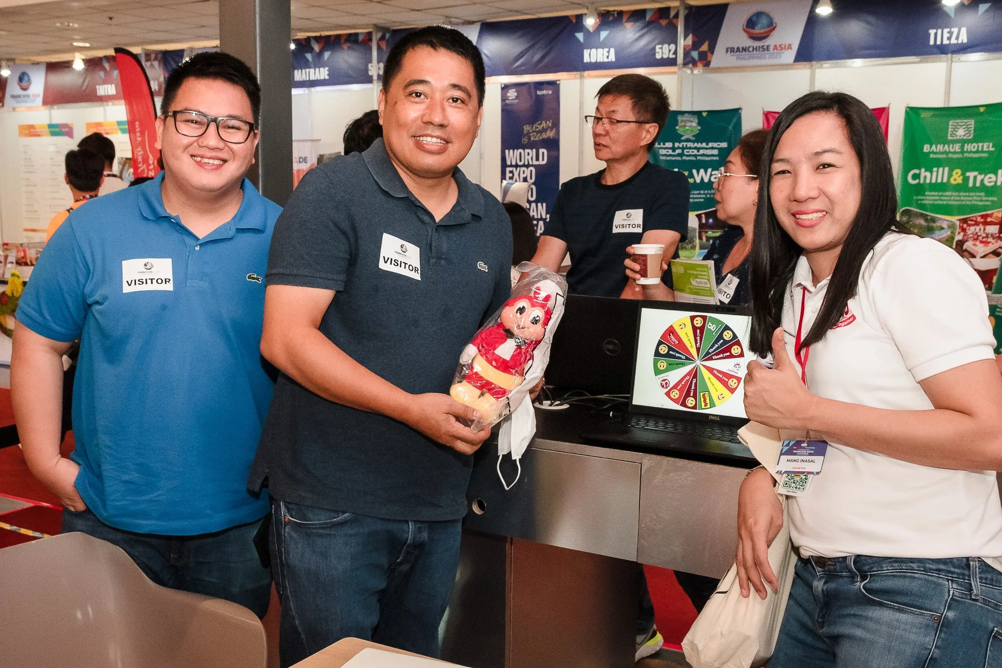 Group of people at an expo, smiling at the camera; one man holds a red plush toy. A woman gives a thumbs-up, and others are standing near a colorful wheel of fortune display and a laptop.