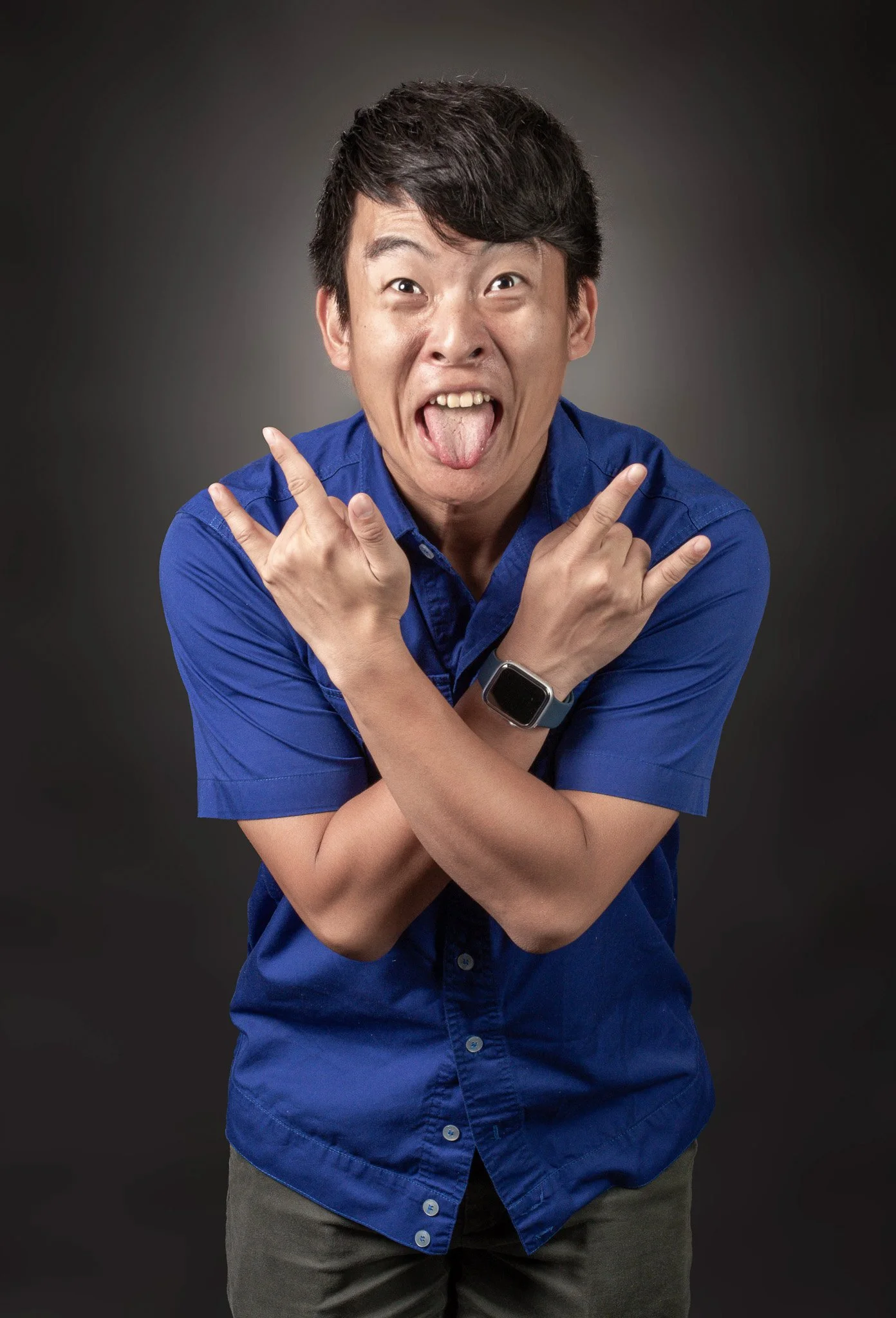 Young man wearing a blue shirt and a smartwatch, making playful facial expressions with tongue out and crossing arms in front, against a dark background.