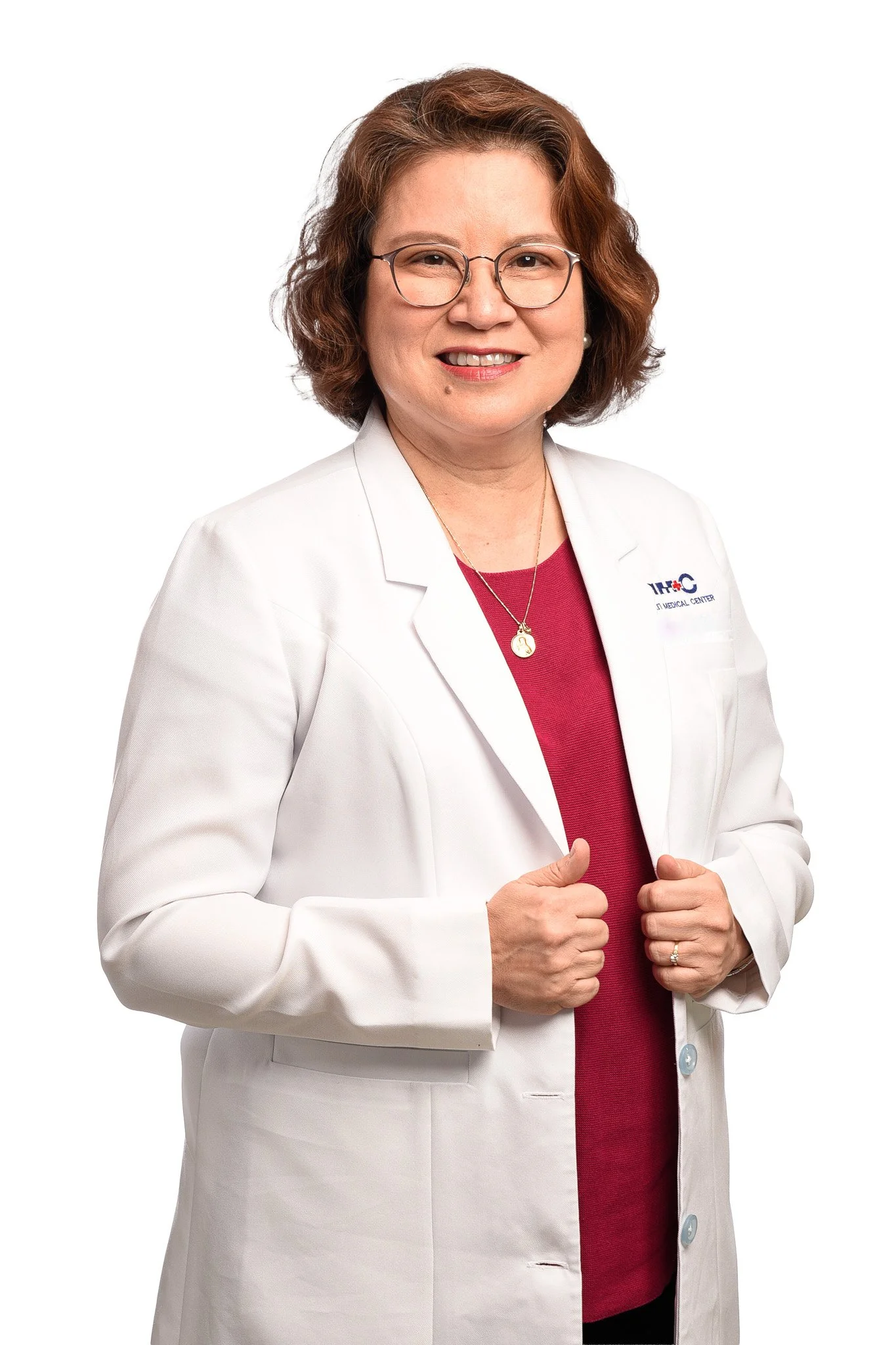 A middle-aged woman with shoulder-length brown hair, wearing glasses, a white medical coat, a red shirt, and a gold necklace, standing against a white background.