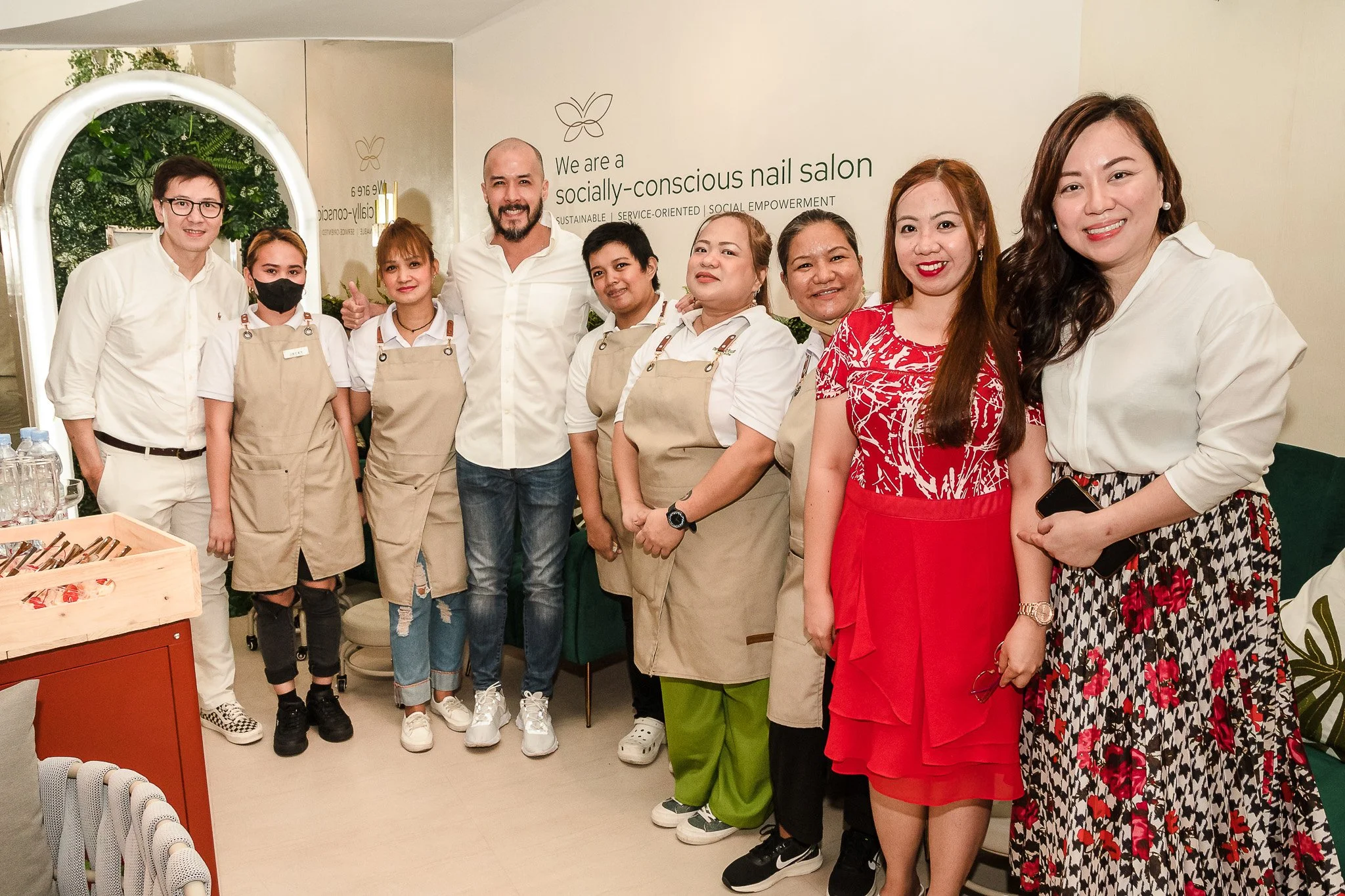 Group of people posing inside a nail salon, with some staff members wearing beige aprons and casual attire, smiling for the photo, with a sign in the background indicating it's a socially-conscious nail salon.