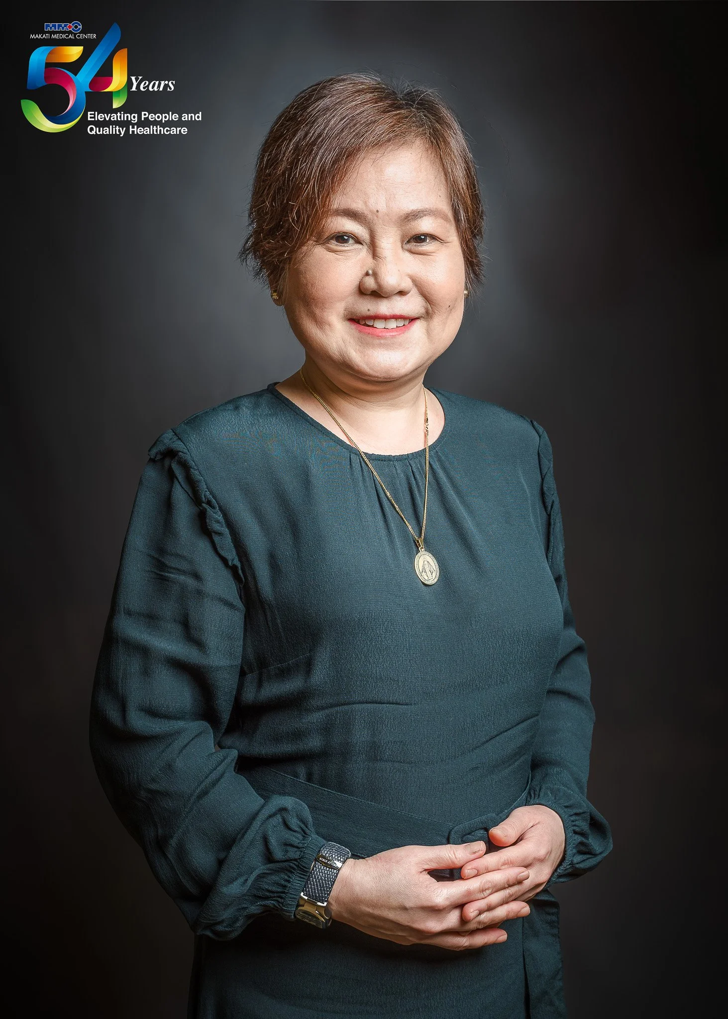 A woman with short brown hair wearing a dark teal dress, jewelry, and a wristwatch, smiling at the camera against a dark background. The top left corner features a logo with the text '54 Years Elevating People and Quality Healthcare.'