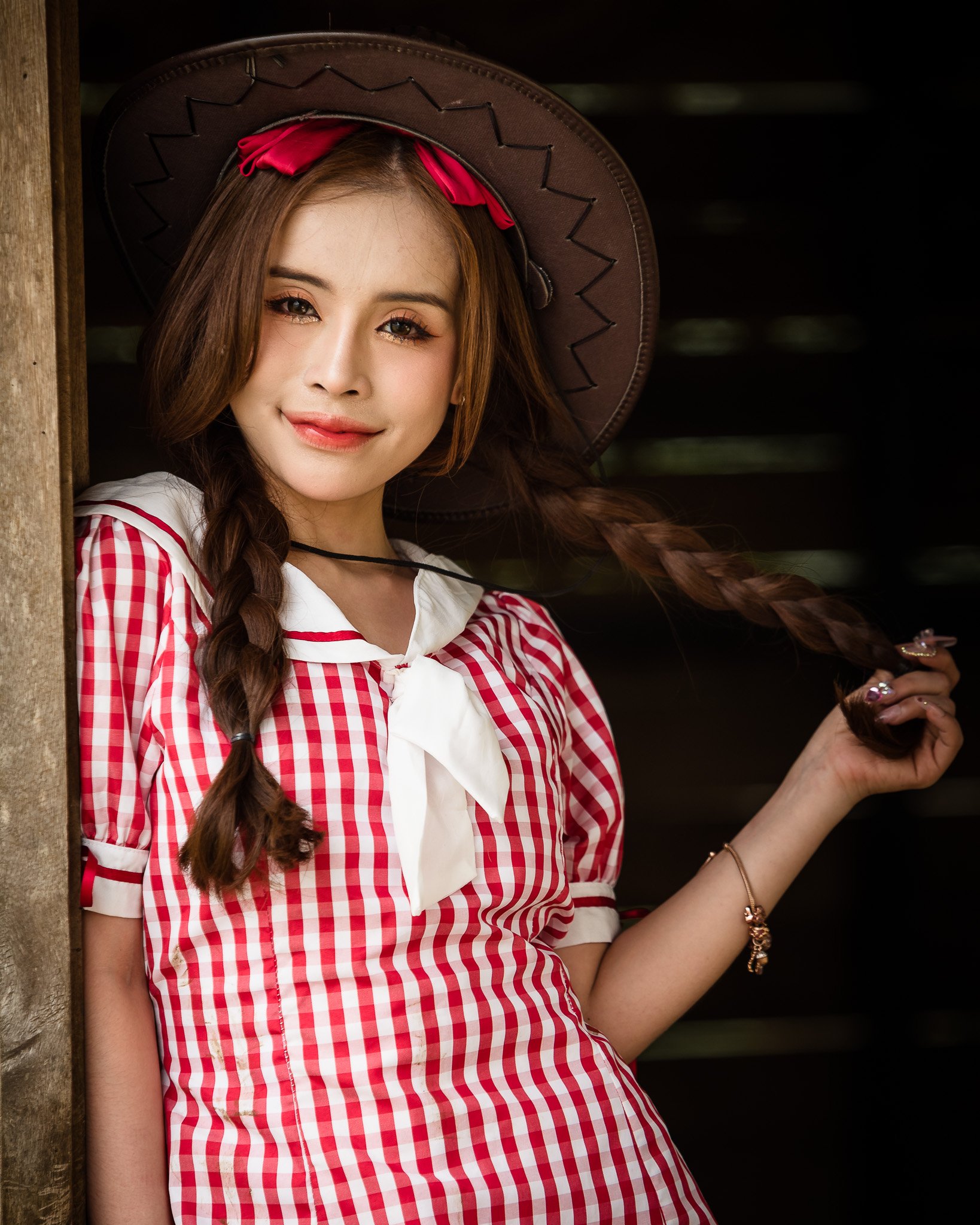 A young woman with braided hair wearing a large cowboy hat, a red and white checkered dress with a white collar and bow, standing next to a wooden structure with a dark background.