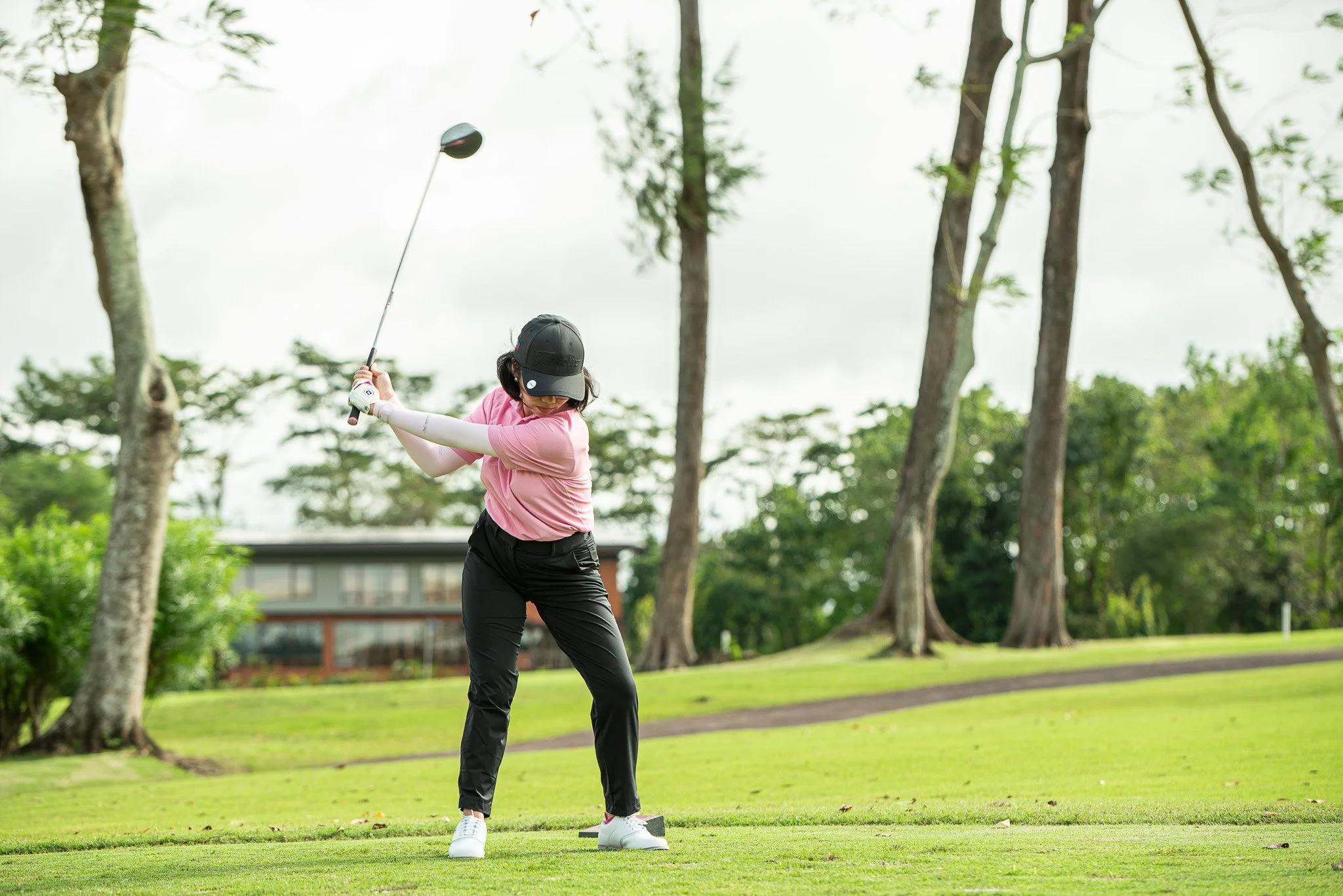 A woman wearing a black cap, pink shirt, black pants, and white shoes is swinging a golf club on a golf course with green grass, trees, and a building in the background.