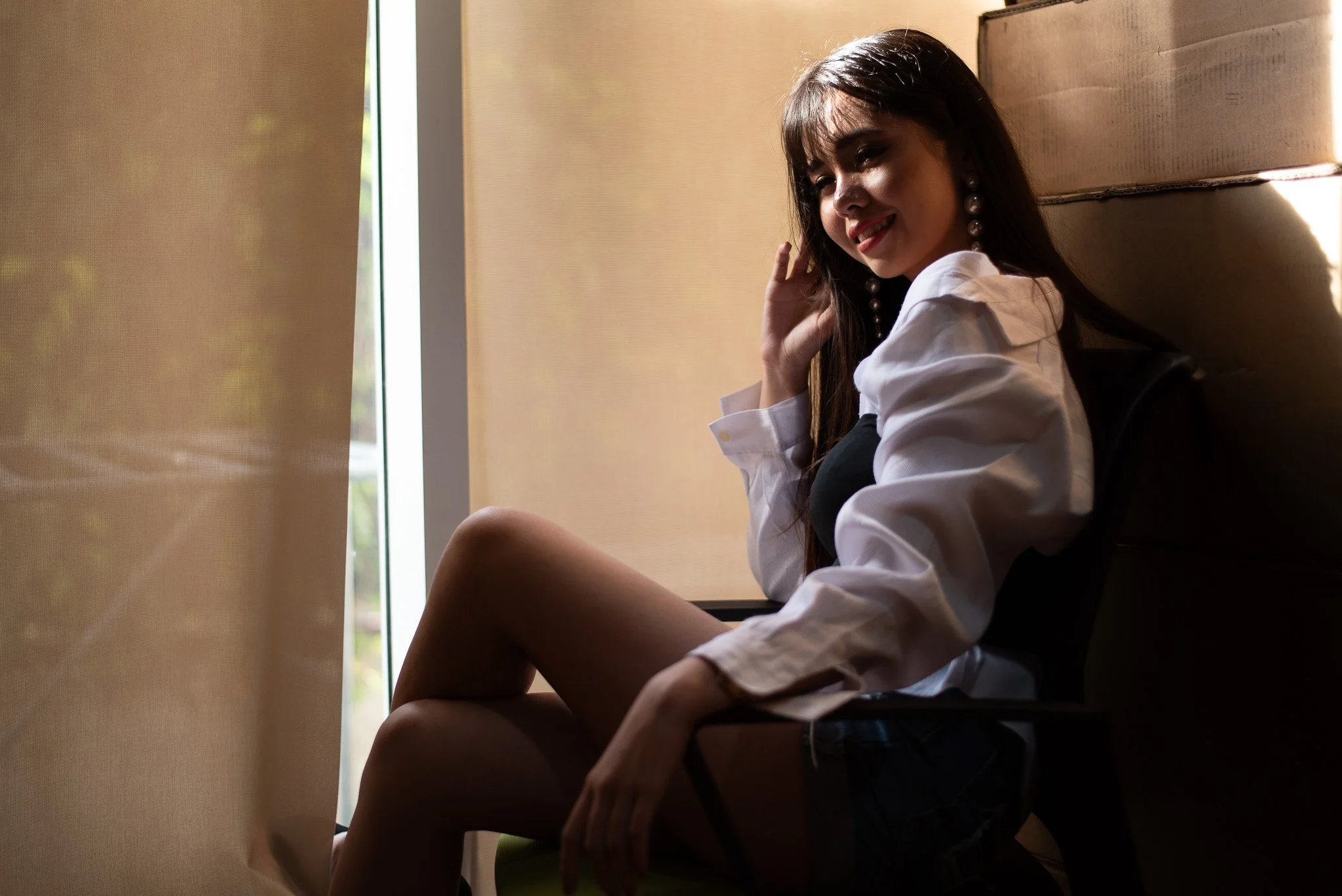 A woman with long dark hair smiling, seated indoors near a window with curtains, wearing a white blouse with rolled-up sleeves and large pearl earrings.