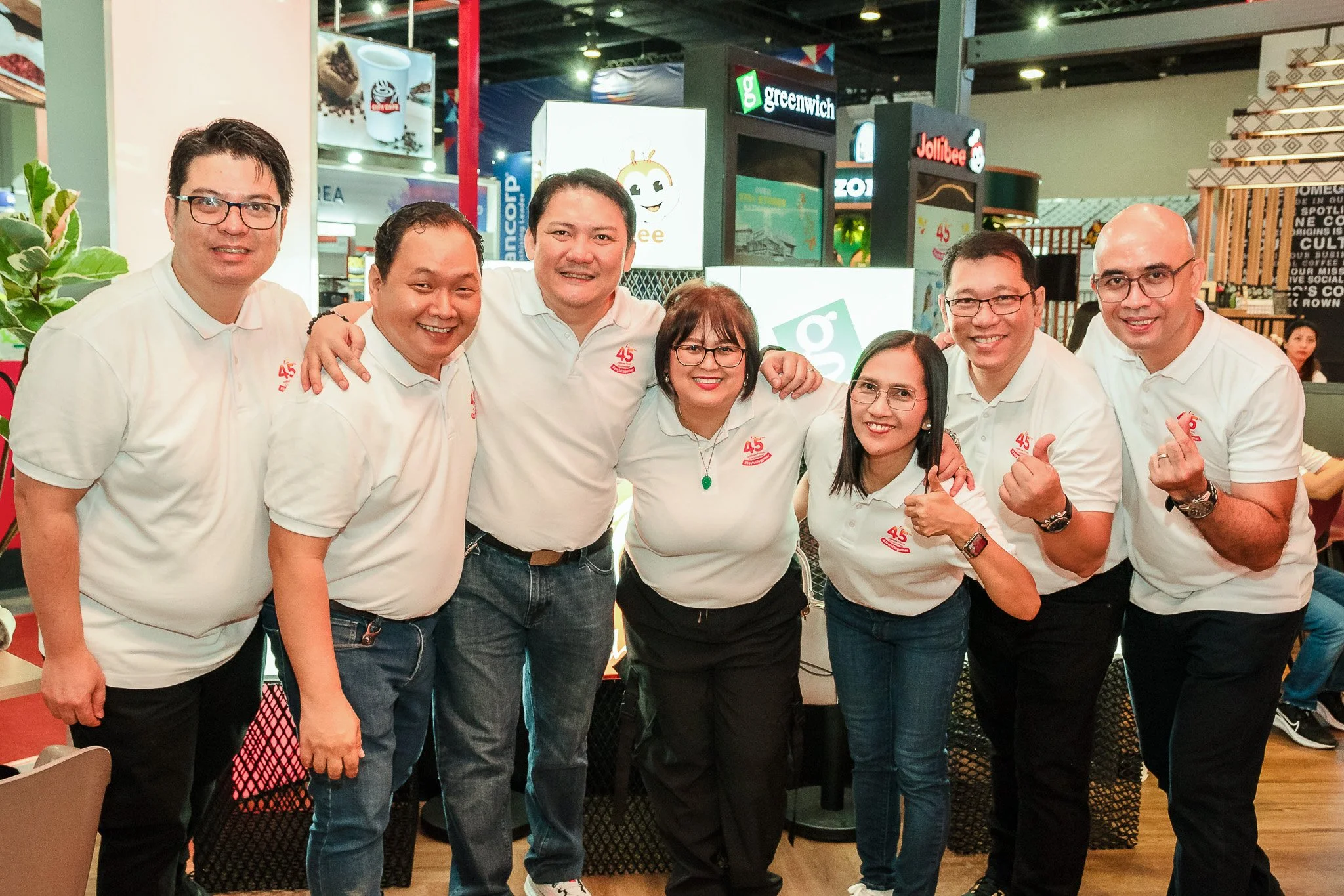 Group of seven smiling people posing at an indoor event, all wearing matching white shirts with a red logo and the number 45. They are standing close together, some with arms around each other, in front of a backdrop with various signs and bright lig
