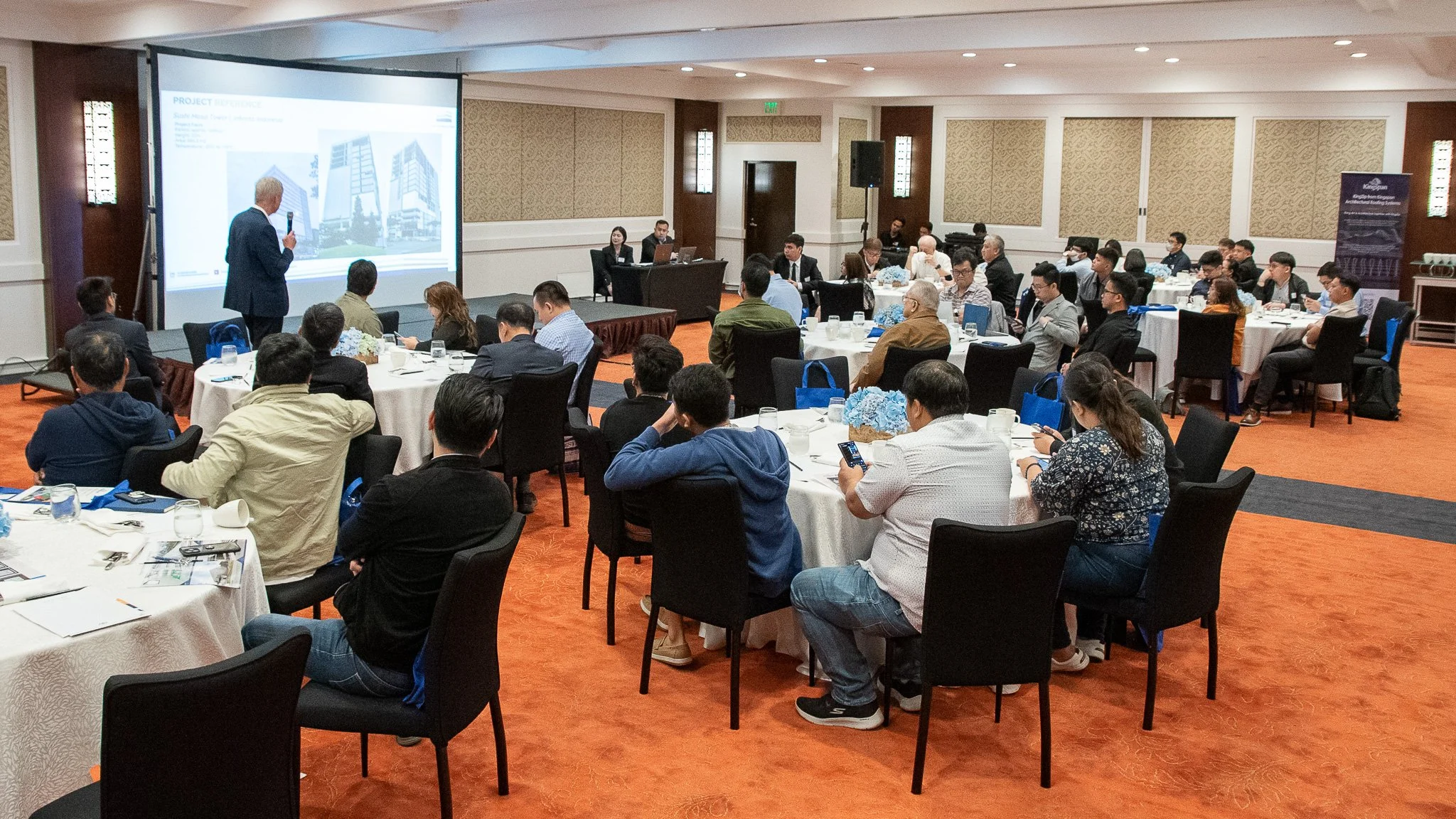A man giving a presentation in a conference room with attendees seated at round tables.