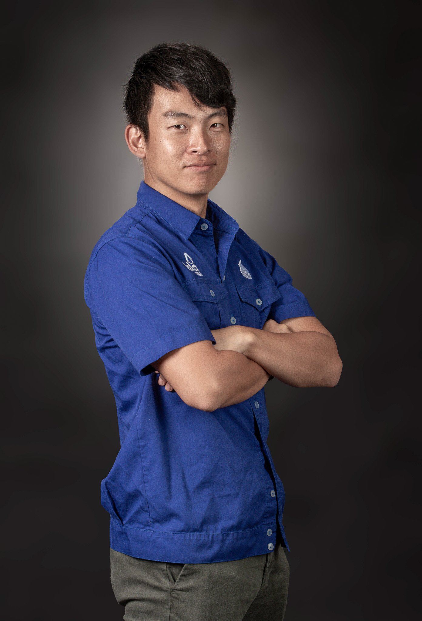 A young man with short black hair is standing against a dark gray background. He is wearing a blue collared shirt with logos on the chest and has his arms crossed, looking confidently at the camera.