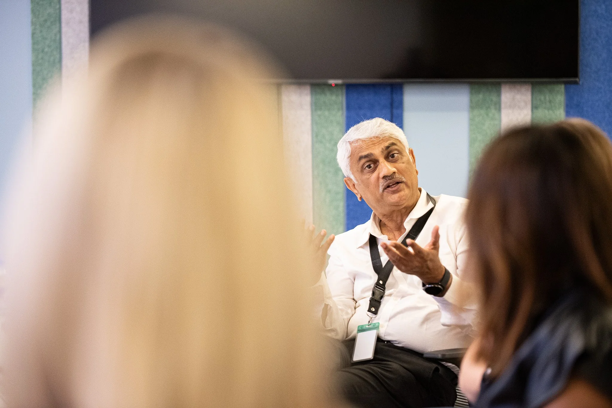 A man with gray hair and a white shirt speaking during a meeting or conference, with people listening, in an office or conference room.