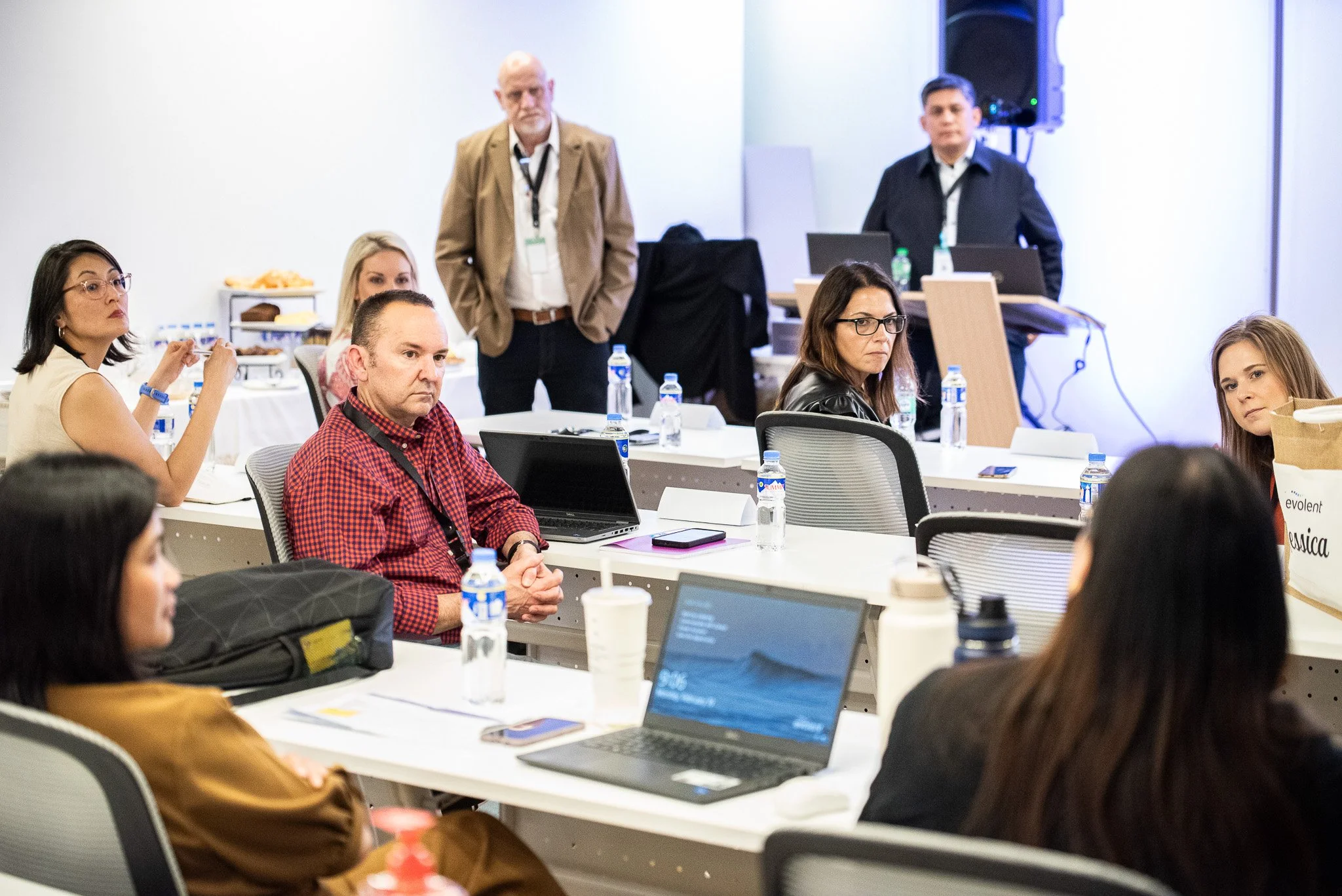 Group of people attending a conference or seminar in a meeting room, sitting at tables with laptops, water bottles, and papers, while two men stand at the back near a speaker and presentation equipment.