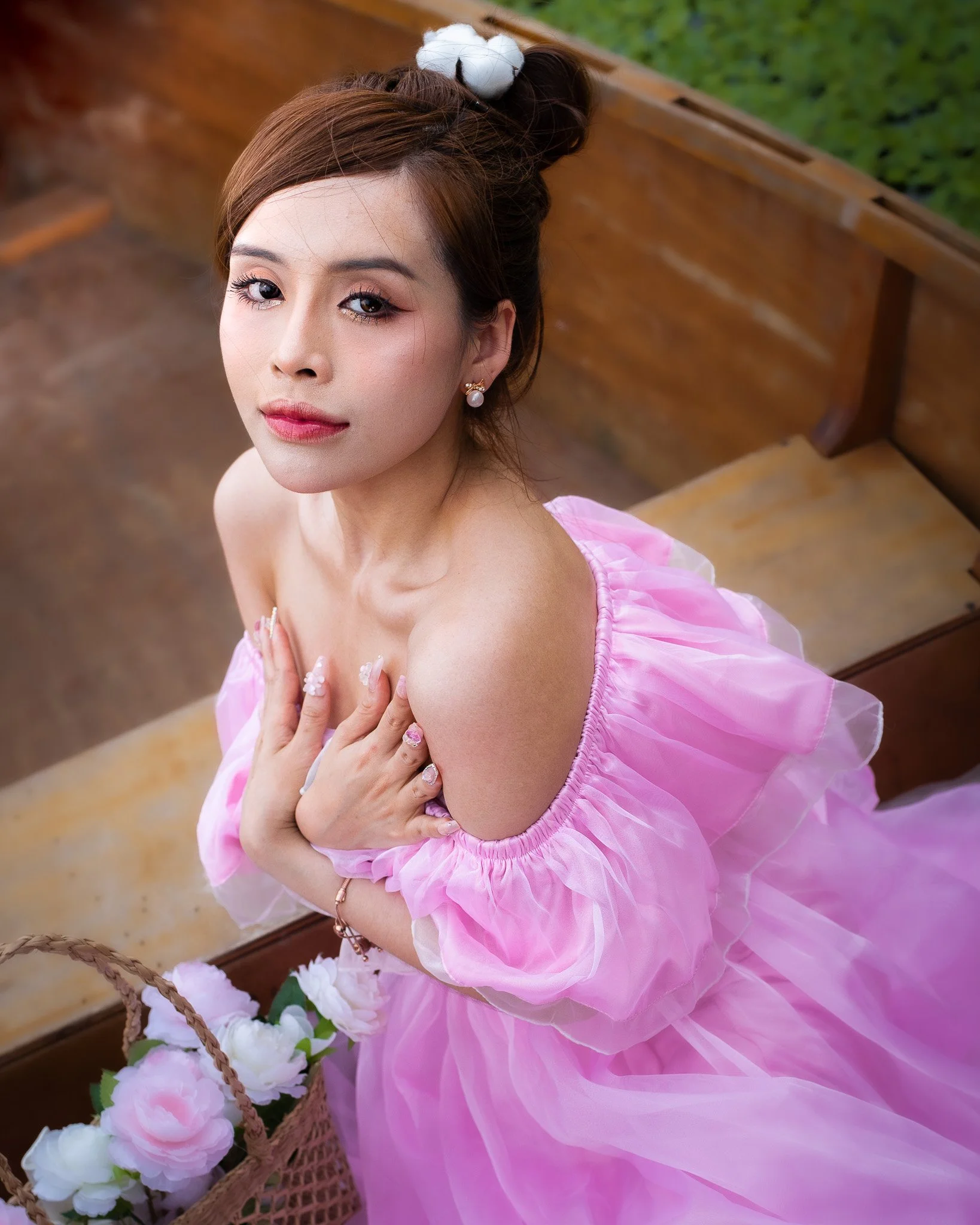A woman in a pink, off-shoulder dress with puffed sleeves sitting on a bench, holding her chest, with a basket of pink and white flowers nearby, looking up at the camera.