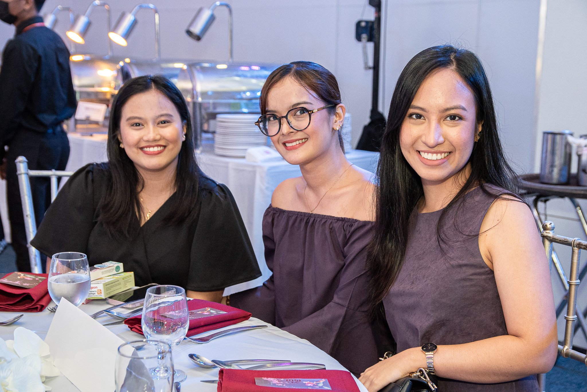 Three women sitting at a formal dinner table, smiling at the camera, with a buffet in the background.