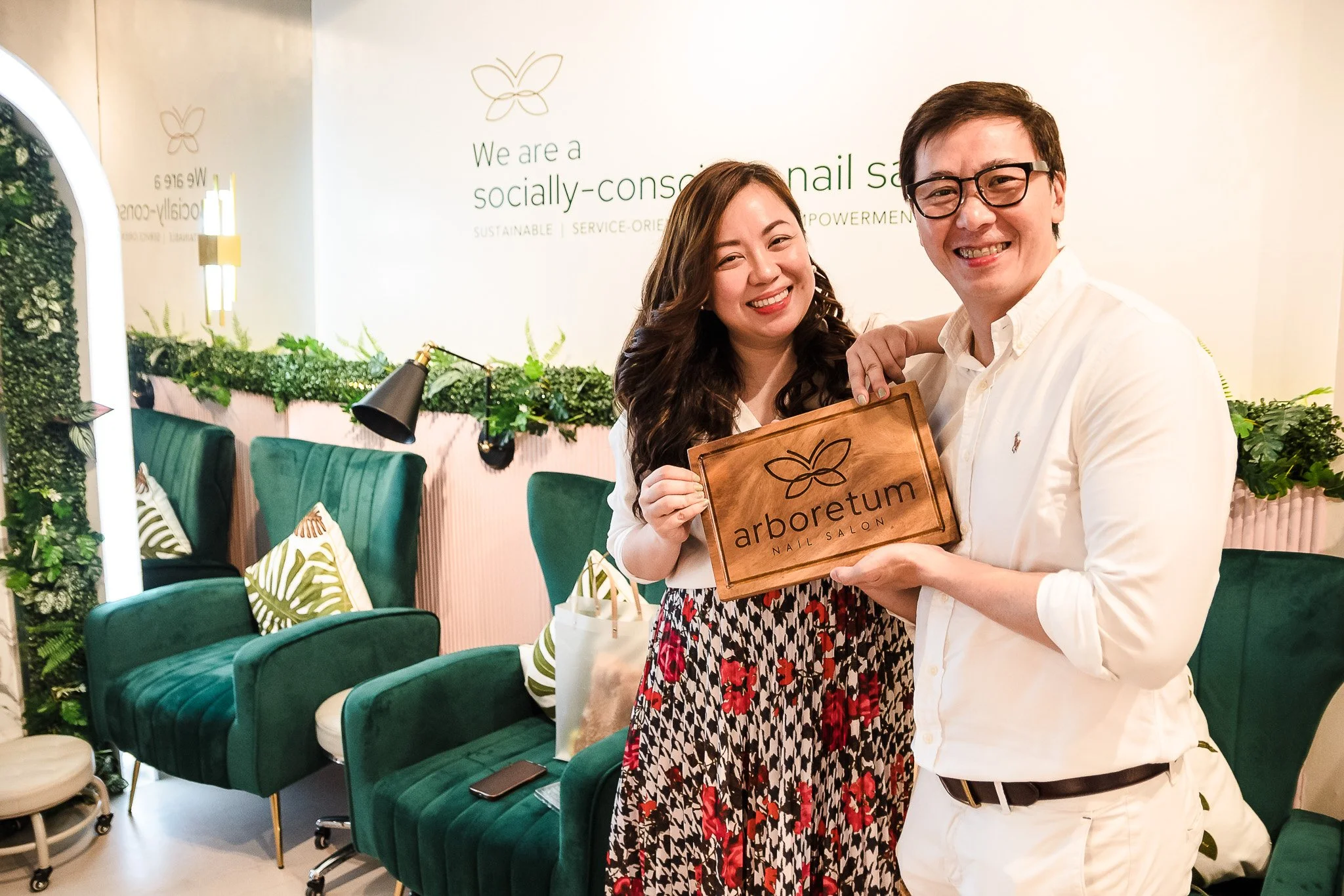 Two women smiling and holding a wooden sign that reads 'arboretum nail salon' inside a nail salon with green chairs and cushions.