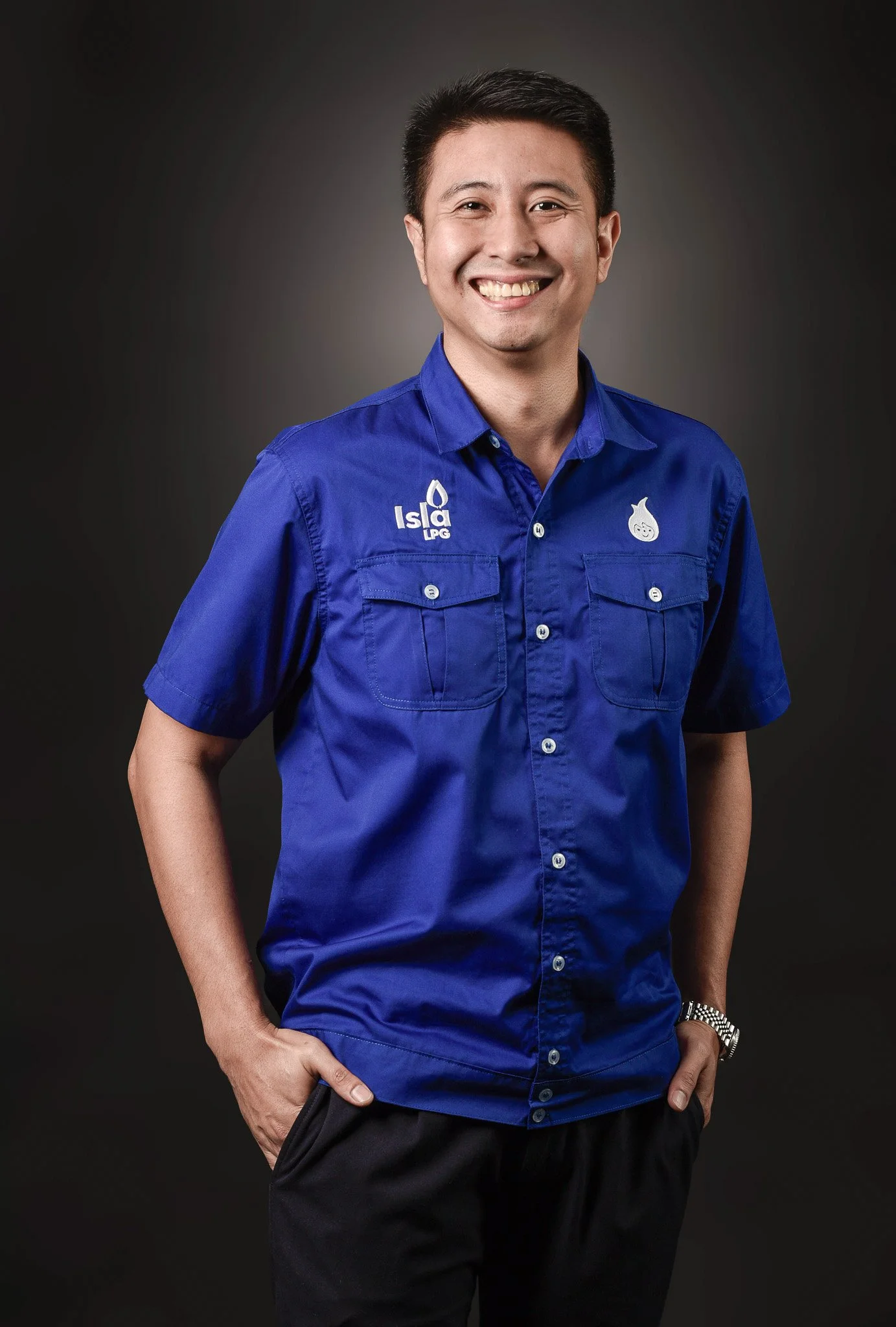 A smiling man with short dark hair wearing a blue work shirt with logos, standing against a dark gray background.