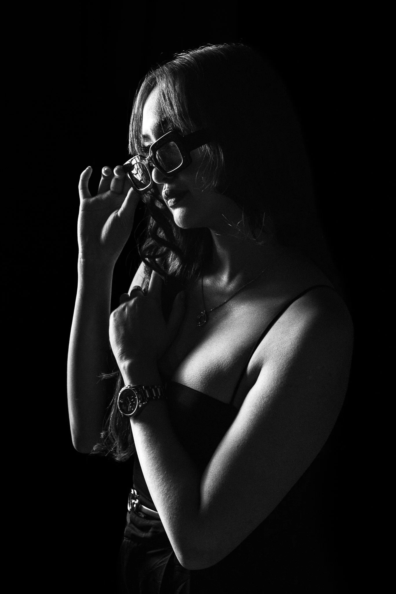 Black and white portrait of a woman with glasses, wearing a watch and jewelry, adjusting her glasses against a dark background.