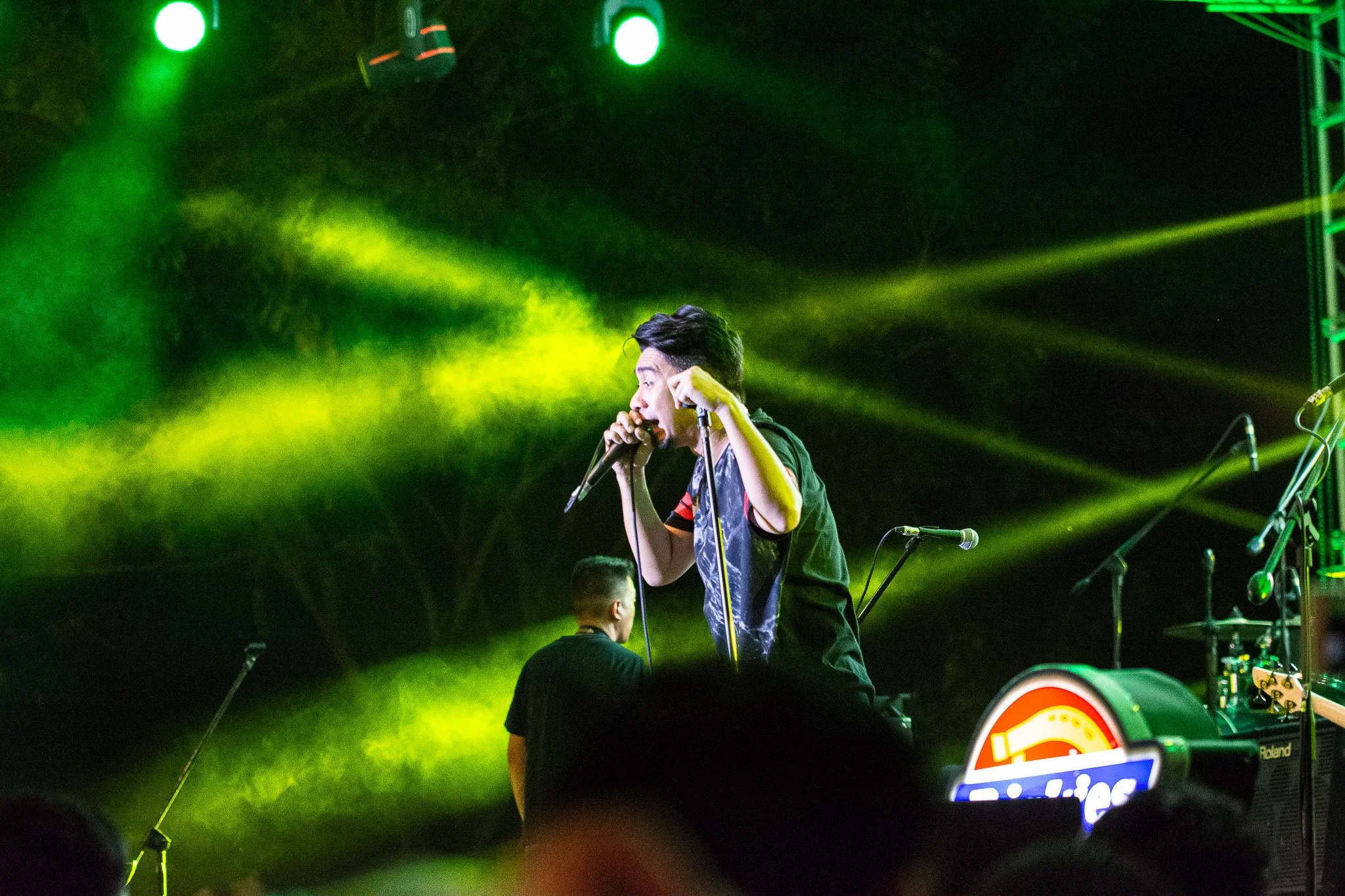 Performer singing on stage with green and yellow stage lights and musical equipment in the background.