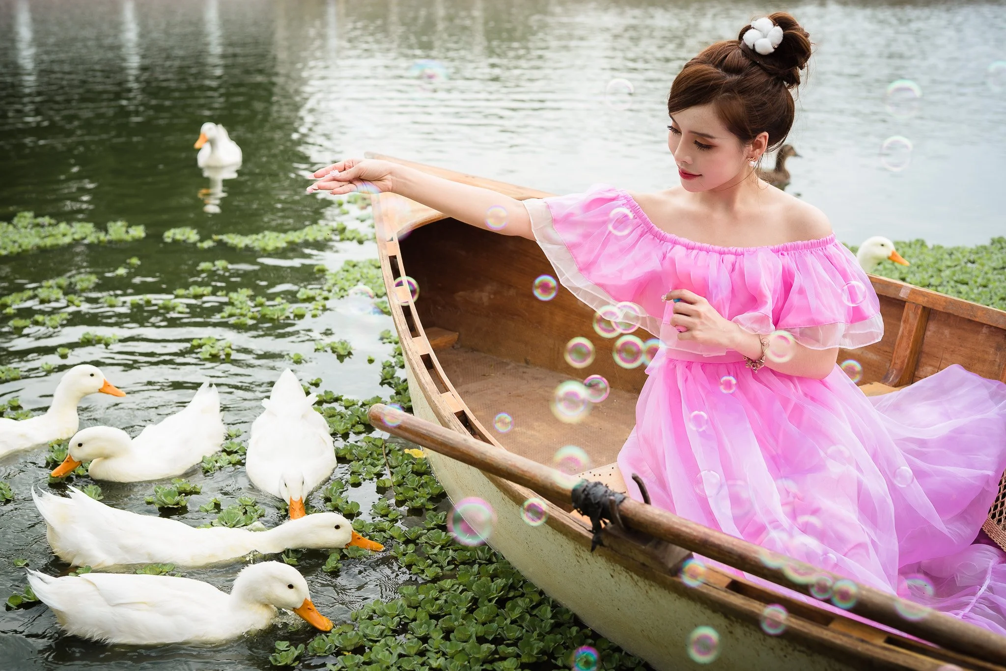 A young woman in a pink dress sitting in a wooden boat on a pond, reaching out to feed a flock of white ducks swimming nearby amidst green lily pads, with soap bubbles floating around.
