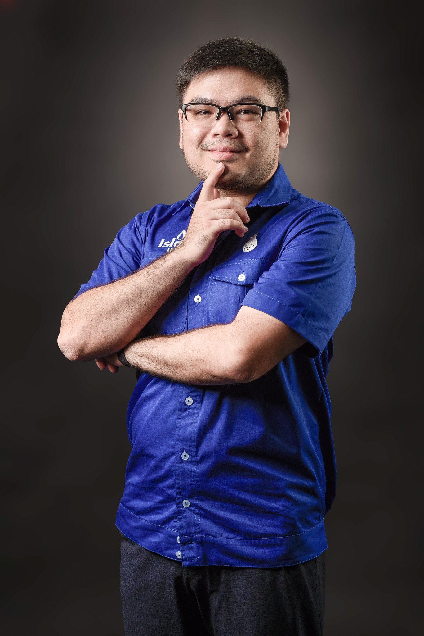 A young man with dark hair and glasses, wearing a blue shirt with a logo, standing with his arms crossed, holding his chin with his right hand, and looking at the camera against a dark background.