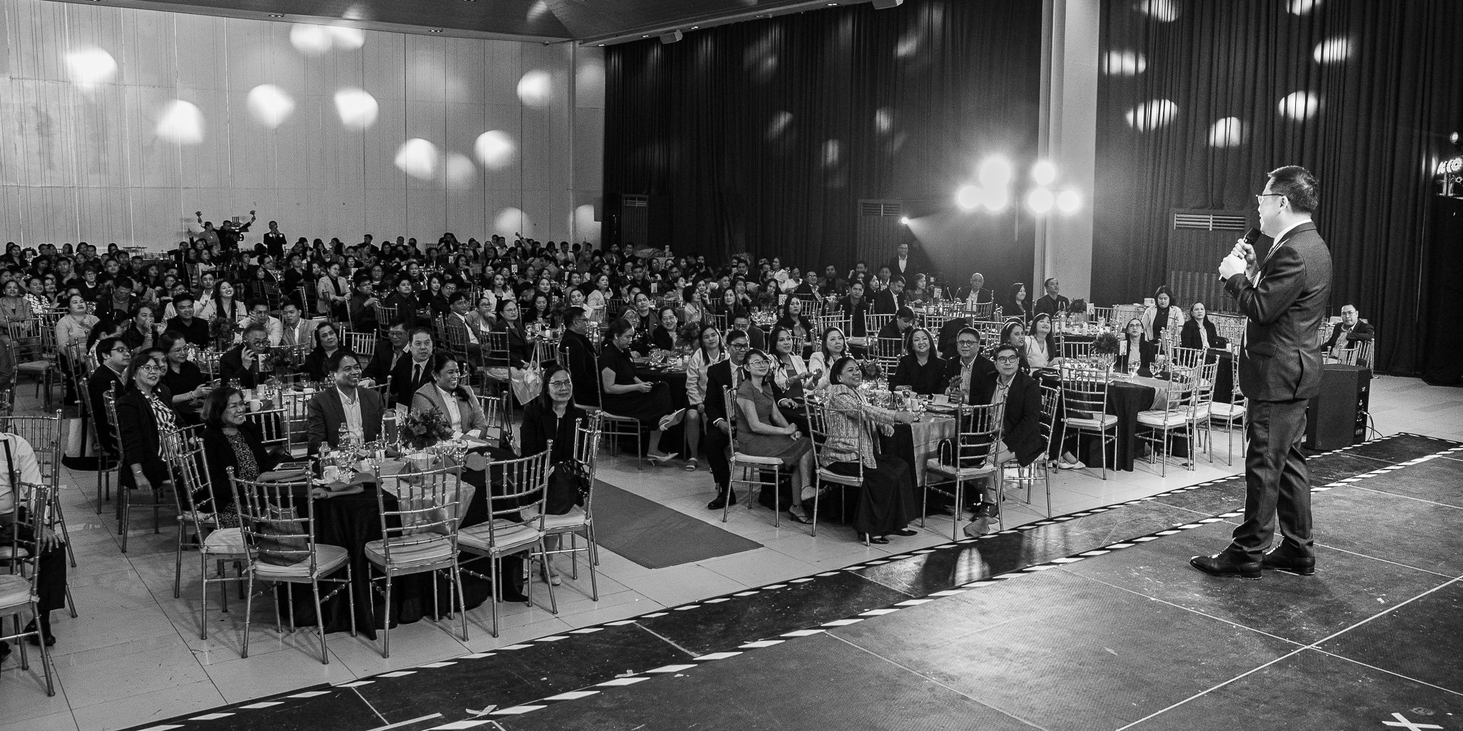 A man in a suit speaking into a microphone on stage at a large formal event, with an audience seated at decorated tables looking towards him.