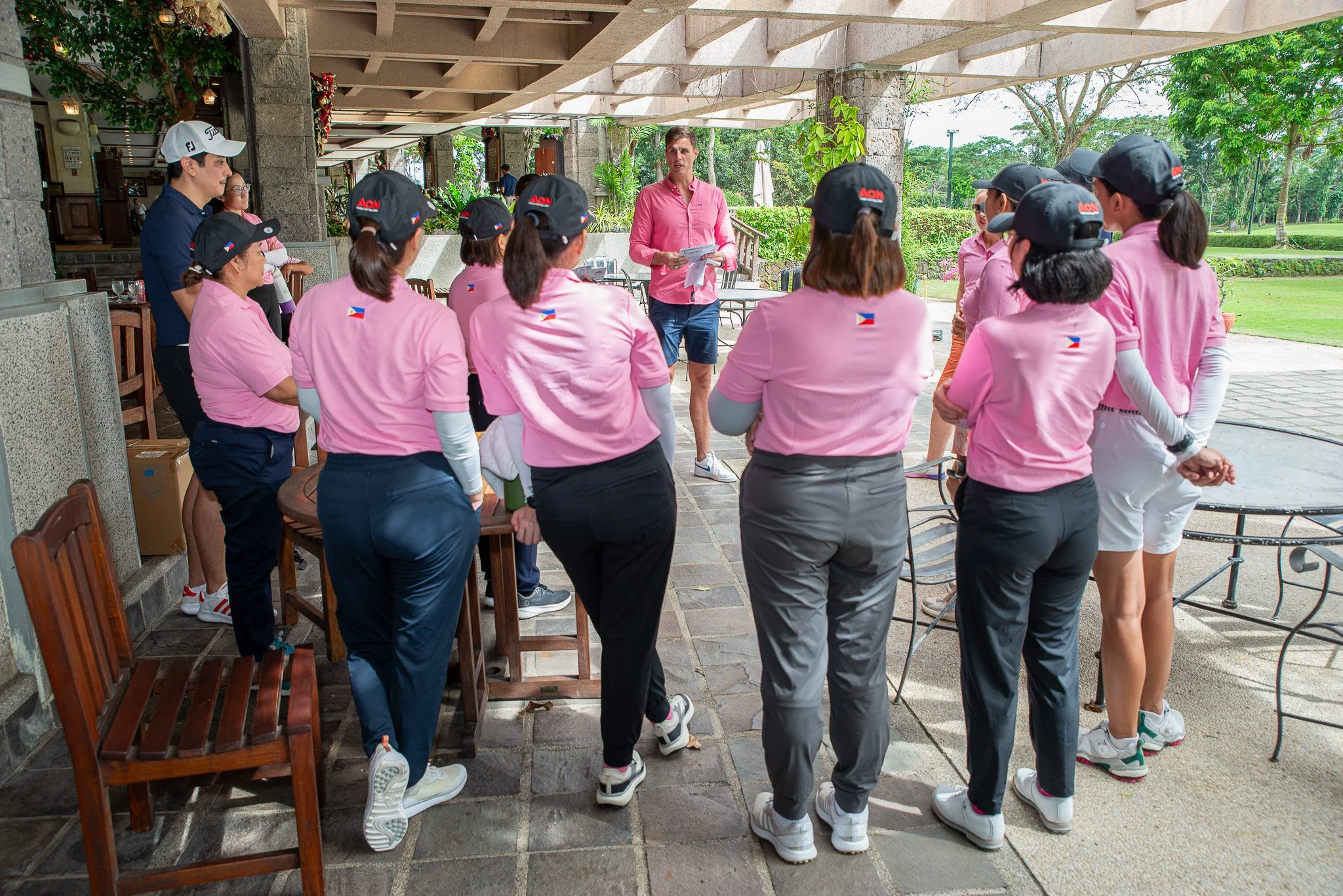 A group of women in pink shirts, some with black hats, listening to a man in a pink shirt who appears to be giving instructions or a briefing outdoors at a golf course or sports club. The women are standing in a semi-circle under a covered patio, and