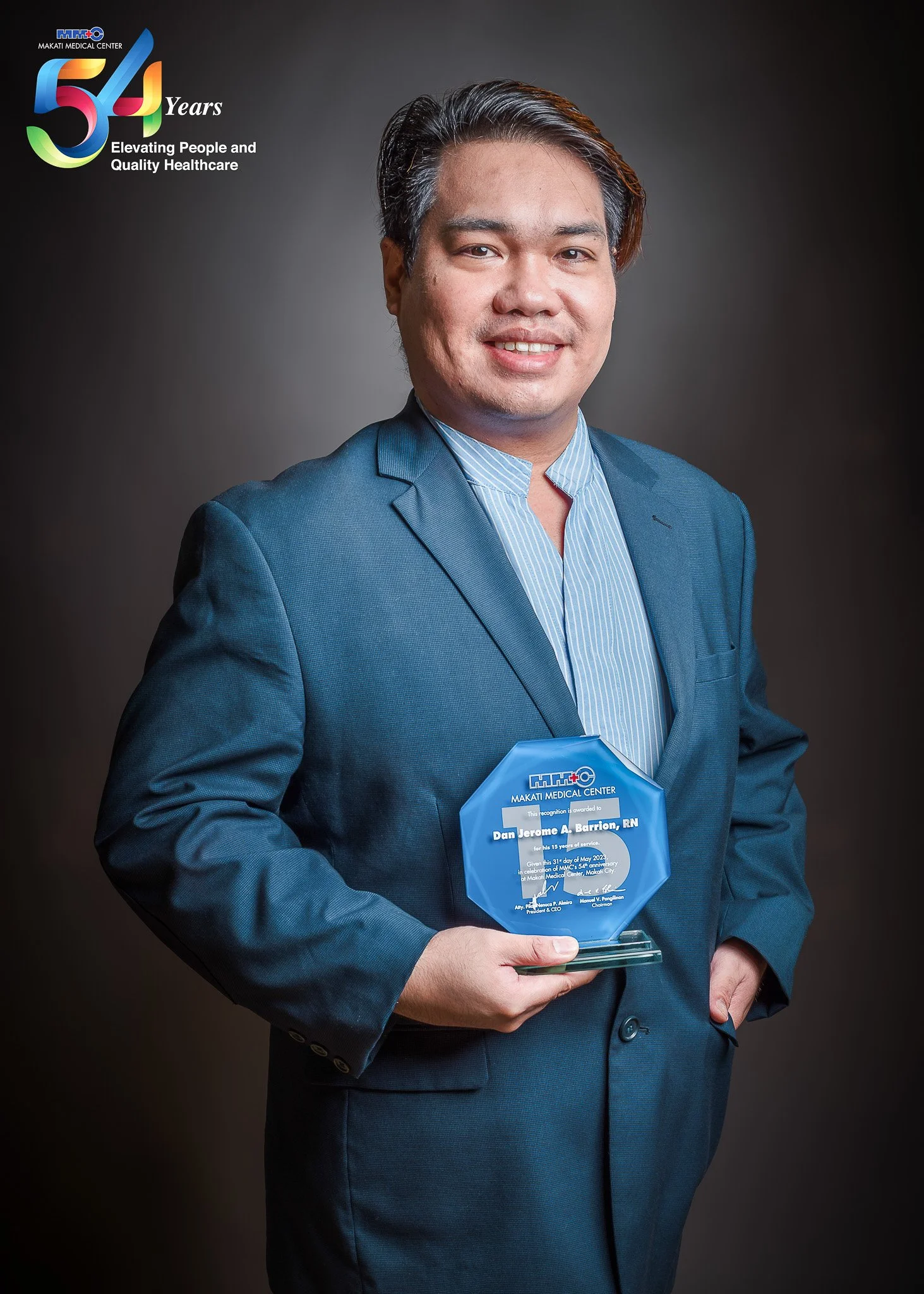 A man in a blue suit holding a glass award in front of a dark background. The award recognizes Jerome A. Barrion, RN from Makati Medical Center for 15 years of service, commemorating their 54th anniversary.