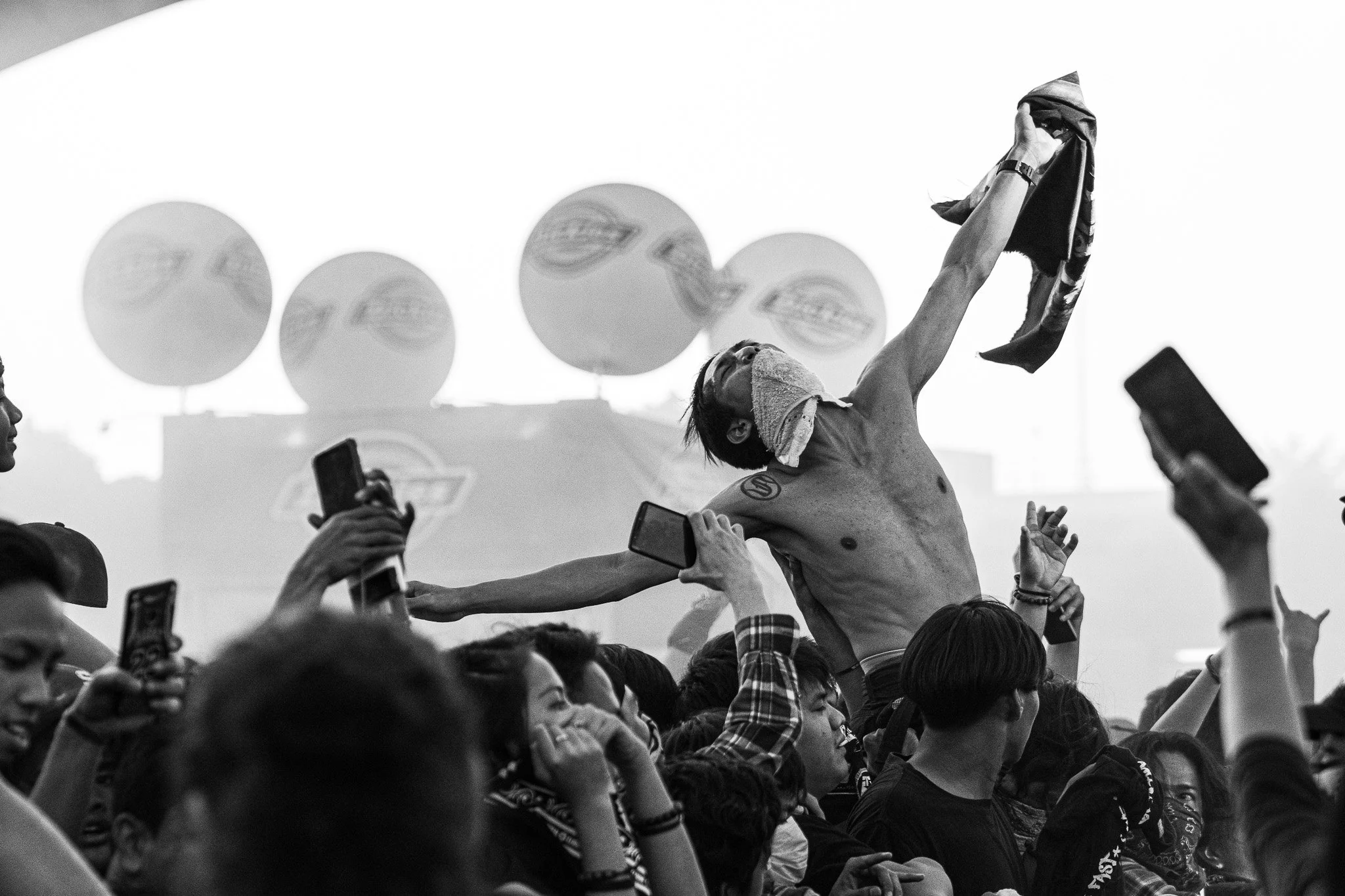 A shirtless man with a towel on his head crowd surfing at a concert, holding a cloth in one hand, surrounded by a large crowd taking photos and cheering, with balloons in the background.