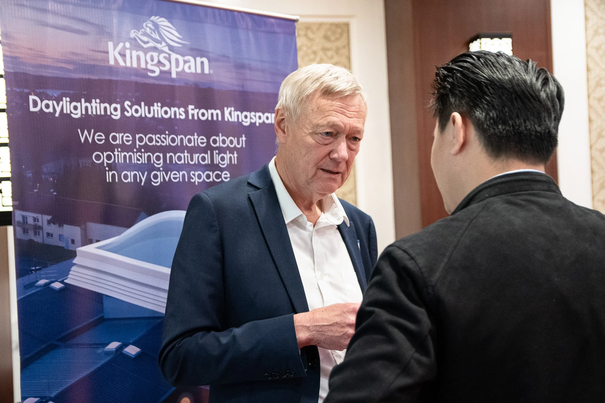 Two men in conversation at a Kingspan booth promoting daylighting solutions, with a large banner in the background displaying the company's name and slogan.
