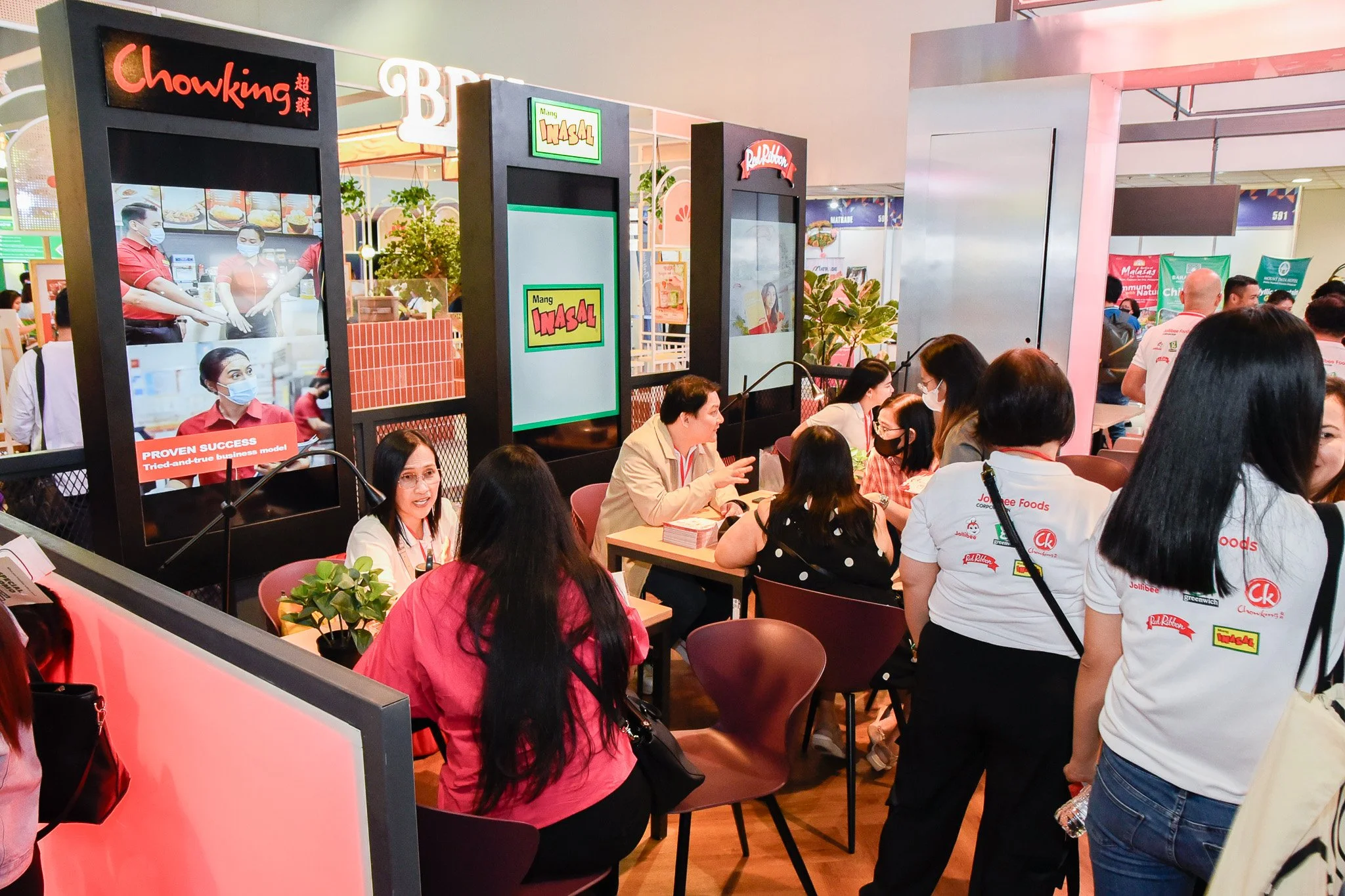 People sitting and standing at a table and booth area, engaging in conversations at a food event or expo. Multiple signs and screens displaying food brand names like Chowking and Mang Inasal are visible.