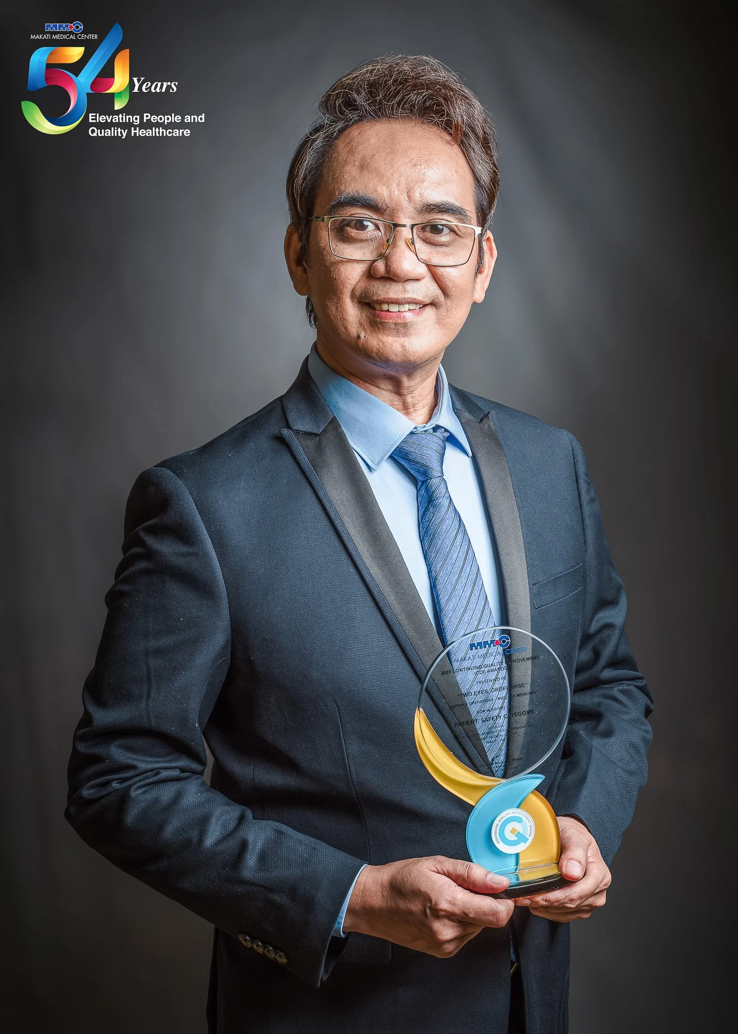 A man dressed in a suit holding a glass award with a yellow and blue design, standing in front of a dark background with a logo and colorful text celebrating 54 years of Makati Medical Center.