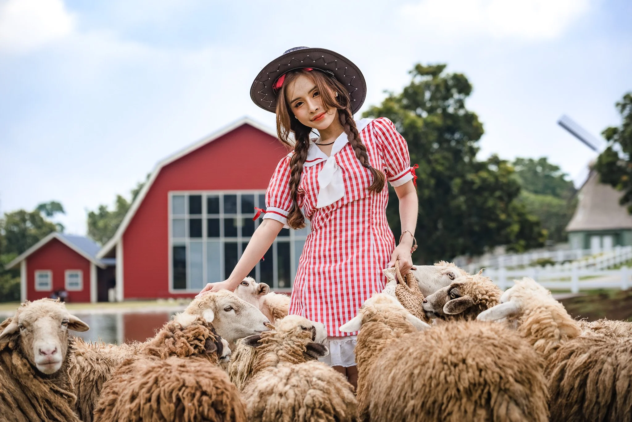 A young woman with braided hair in a red and white checkered dress and a wide-brimmed hat standing among sheep with a red barn and farm buildings in the background.