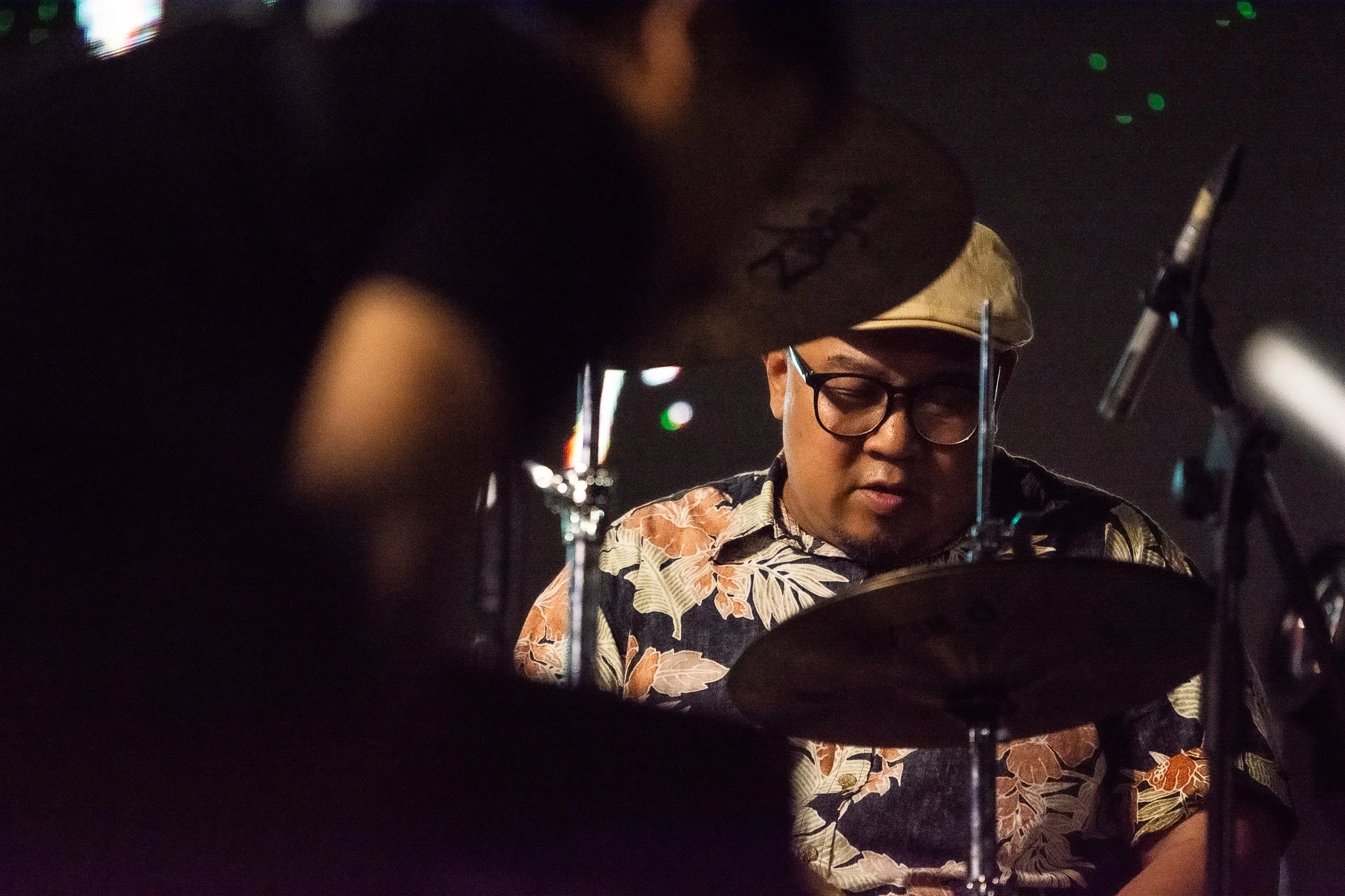 A man with glasses wearing a floral patterned shirt and a beige cap, playing drums in a dimly lit setting.