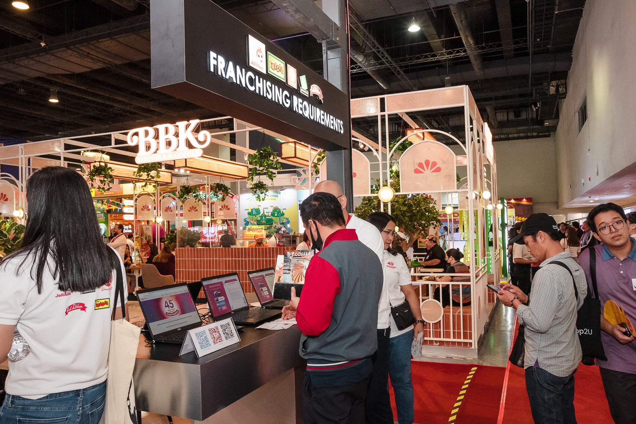 People standing at a registration or check-in counter at a trade show or exhibition, with booths and signage in the background.