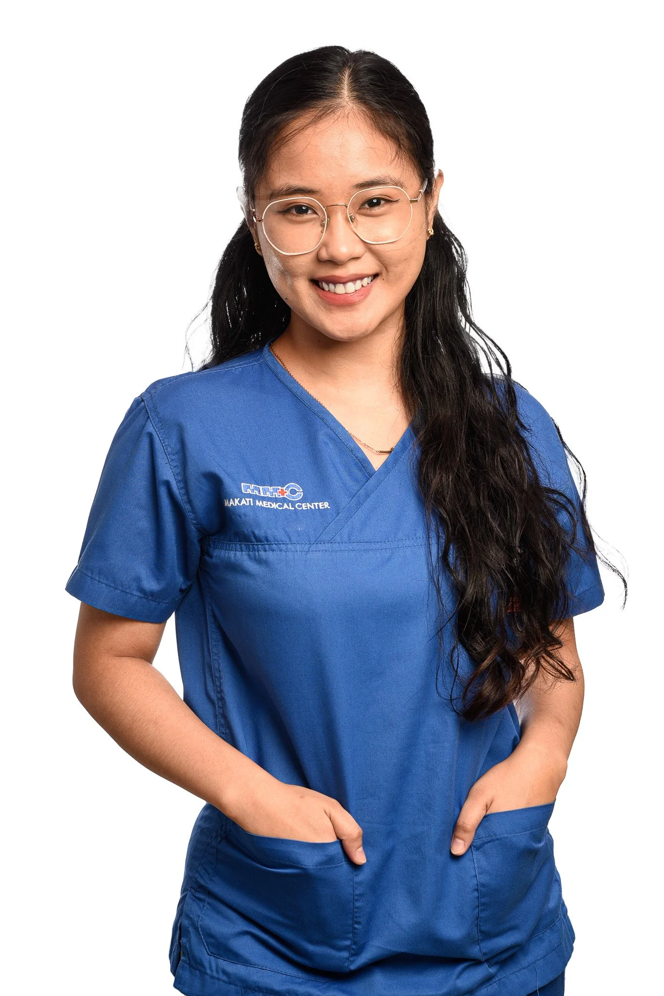 A young woman wearing blue scrubs with her hands in her pockets, smiling at the camera. The scrubs have a logo for Makati Medical Center.