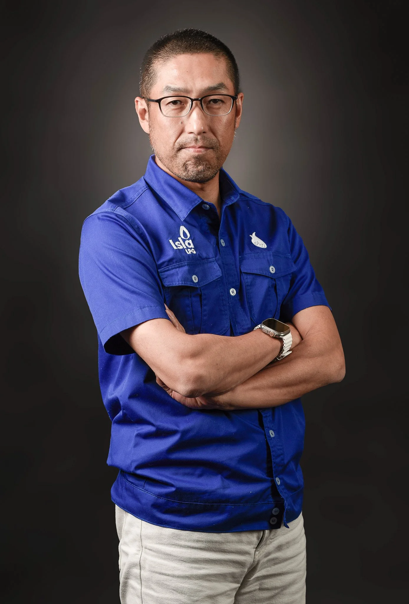 Man with glasses and short hair, wearing a blue work shirt and watch, standing with arms crossed against a dark gray background.
