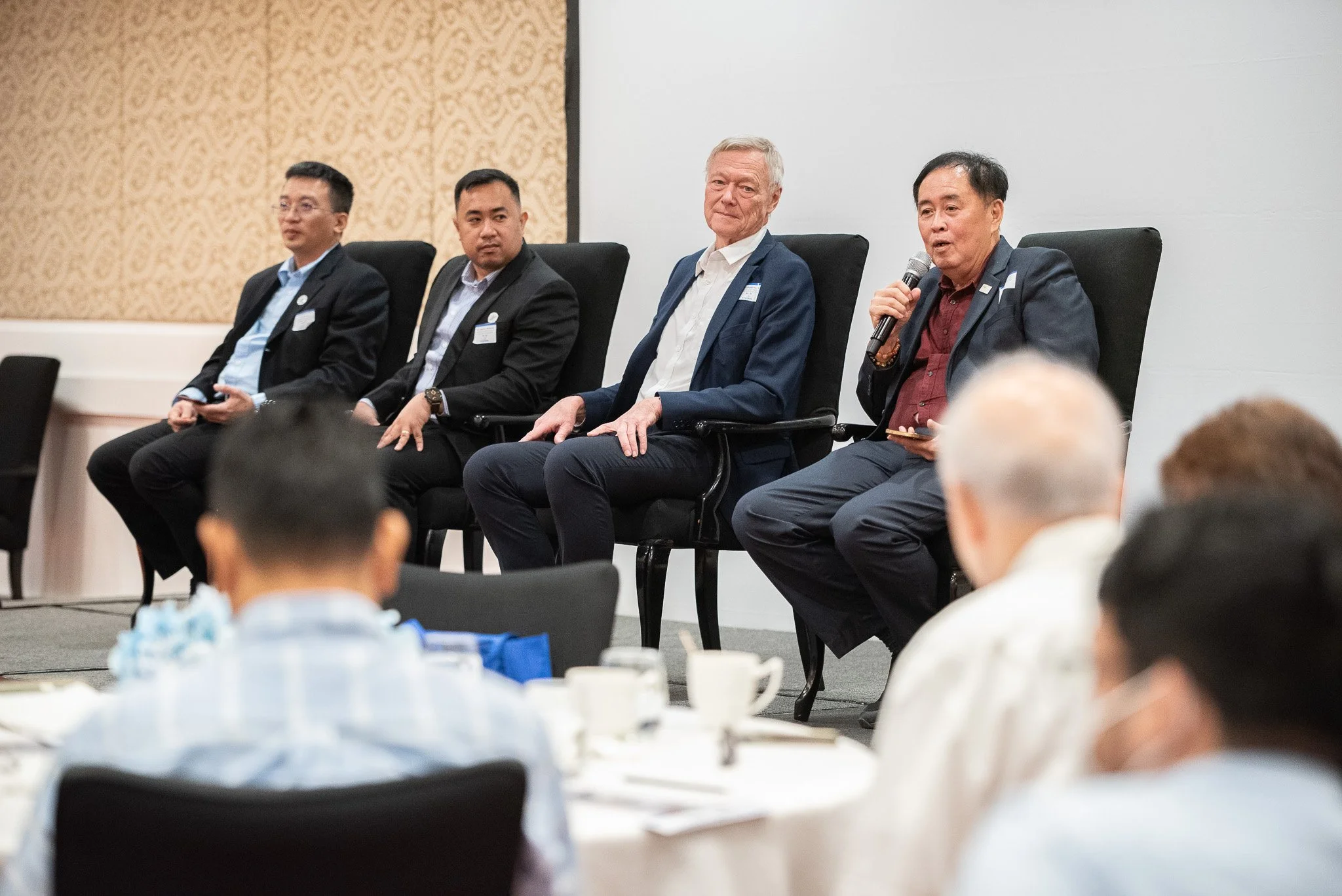 A panel of five men in suits seated on a stage, one man speaking into a microphone. Audience members are visible in the foreground.