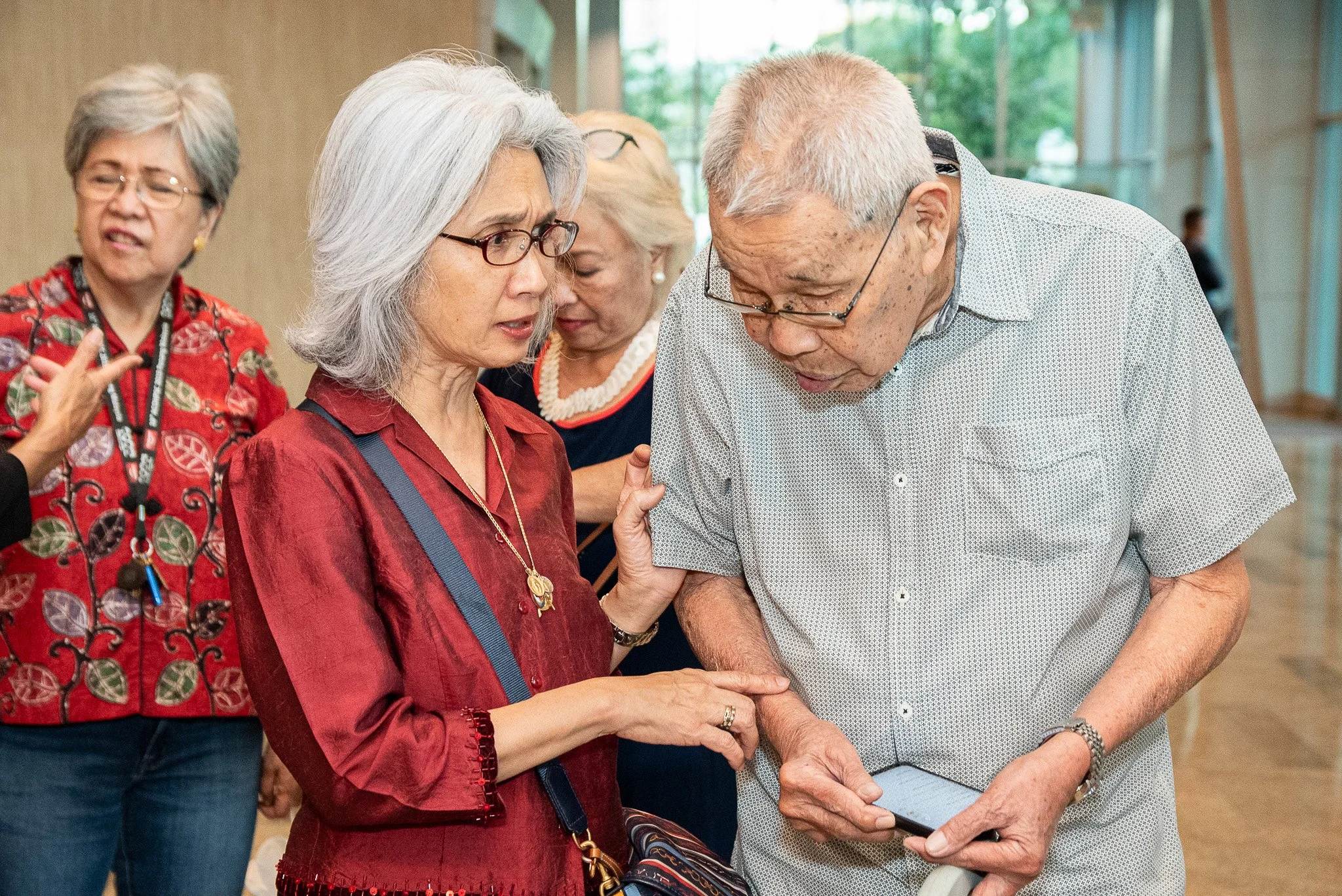 A group of elderly Asian people gathered in a room, with one woman showing something on her phone to a man, while others look on.
