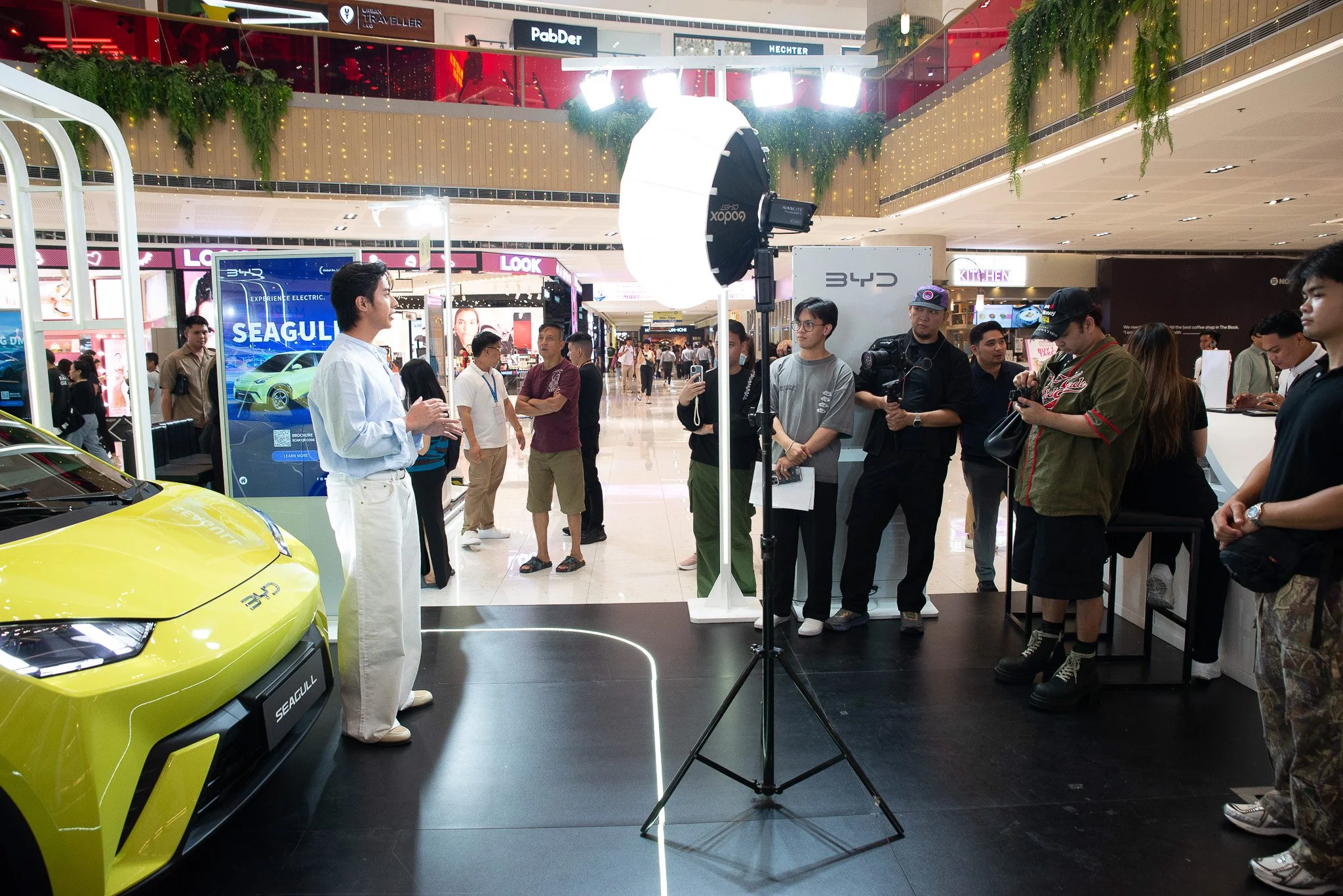 A person is giving a presentation or interview at an event in a shopping mall, with a yellow car and a group of people and photographers standing nearby. There are bright lights, a large screen displaying a car, and mall signage in the background.