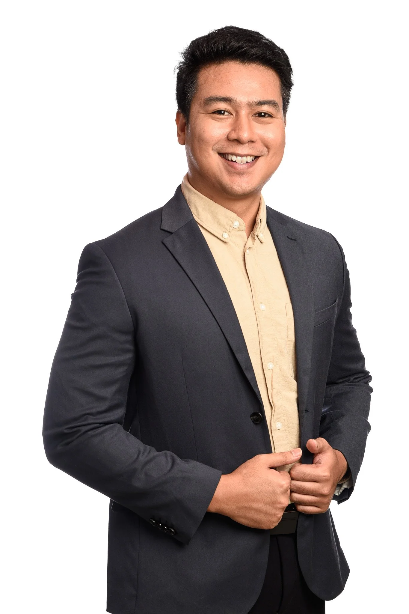 A young man in a dark blazer and beige dress shirt smiling and looking at the camera, with his hands adjusting his blazer.