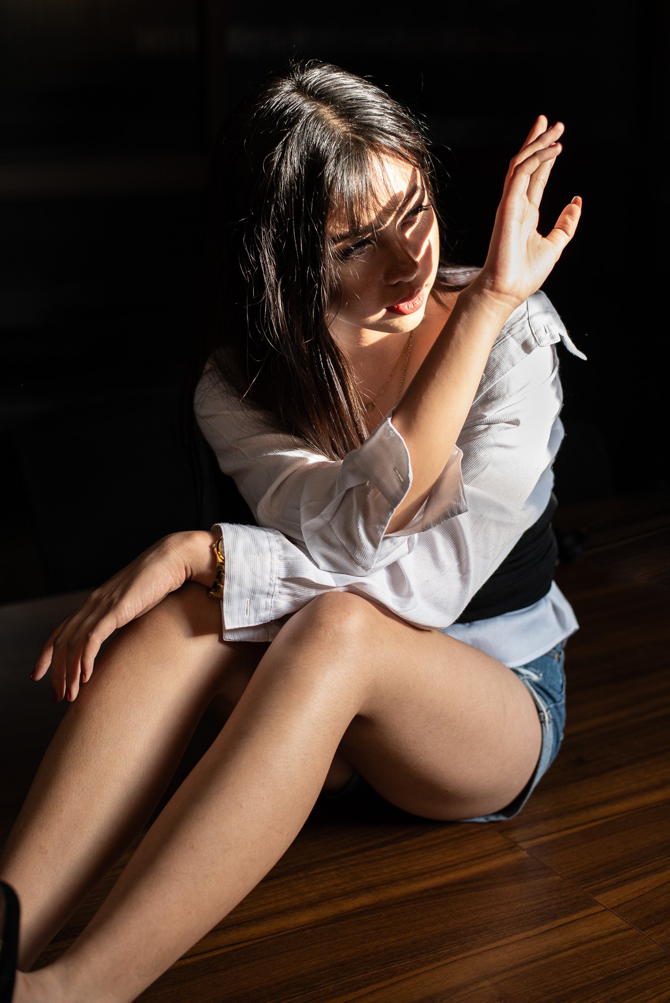 A woman with long dark hair sitting on a wooden floor, wearing a white shirt and denim shorts, with sunlight highlighting her face and upper body, creating shadows.