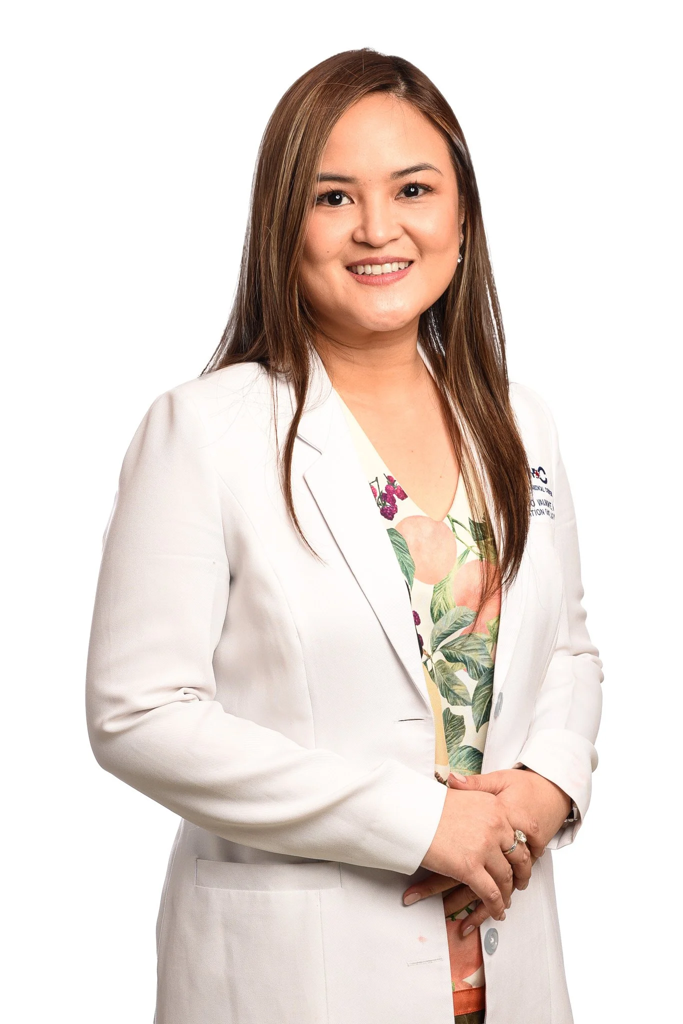 A woman in a white medical coat standing and smiling, with long brown hair, against a white background.